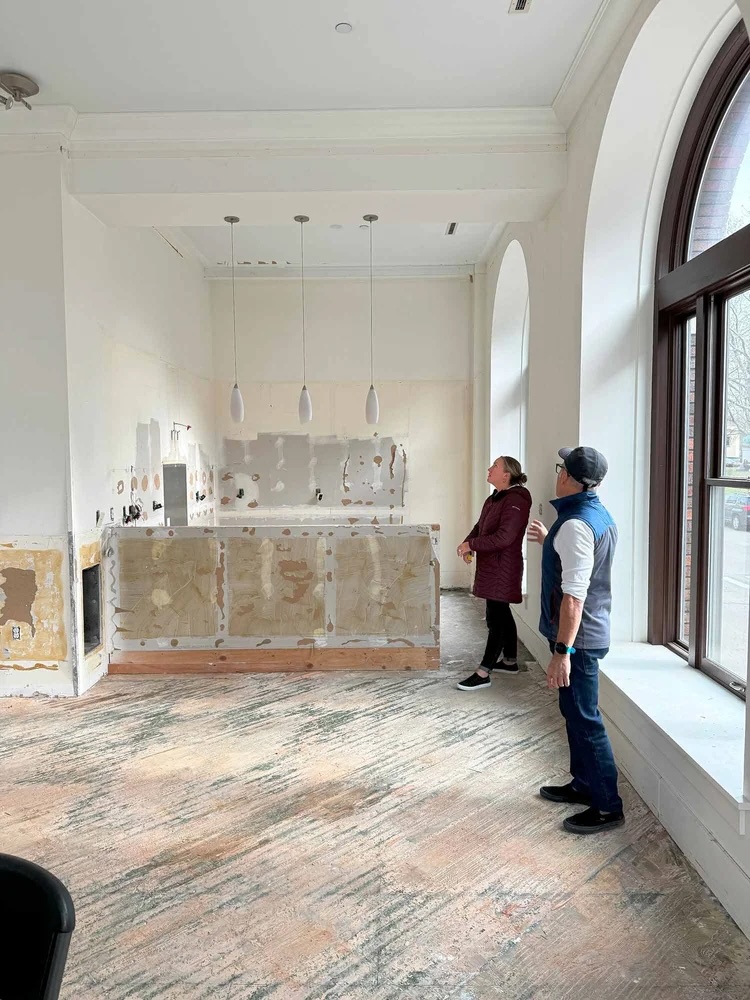 Two people stand in an unfinished room with bare walls and markings, near large arched windows.