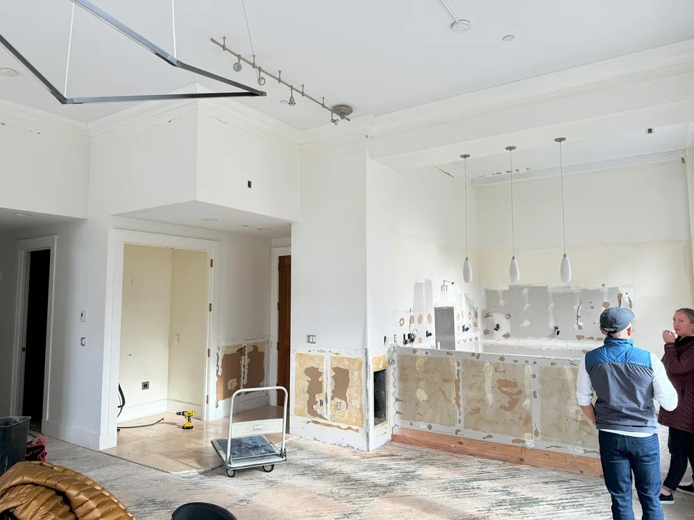 Two people standing in a partially renovated white kitchen with missing cabinets and exposed wall patches.