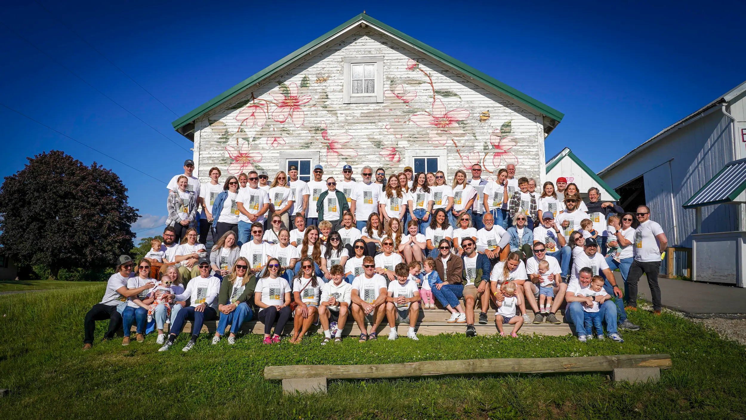Large group of people wearing matching white shirts posing outside a flower-painted barn on green grass.