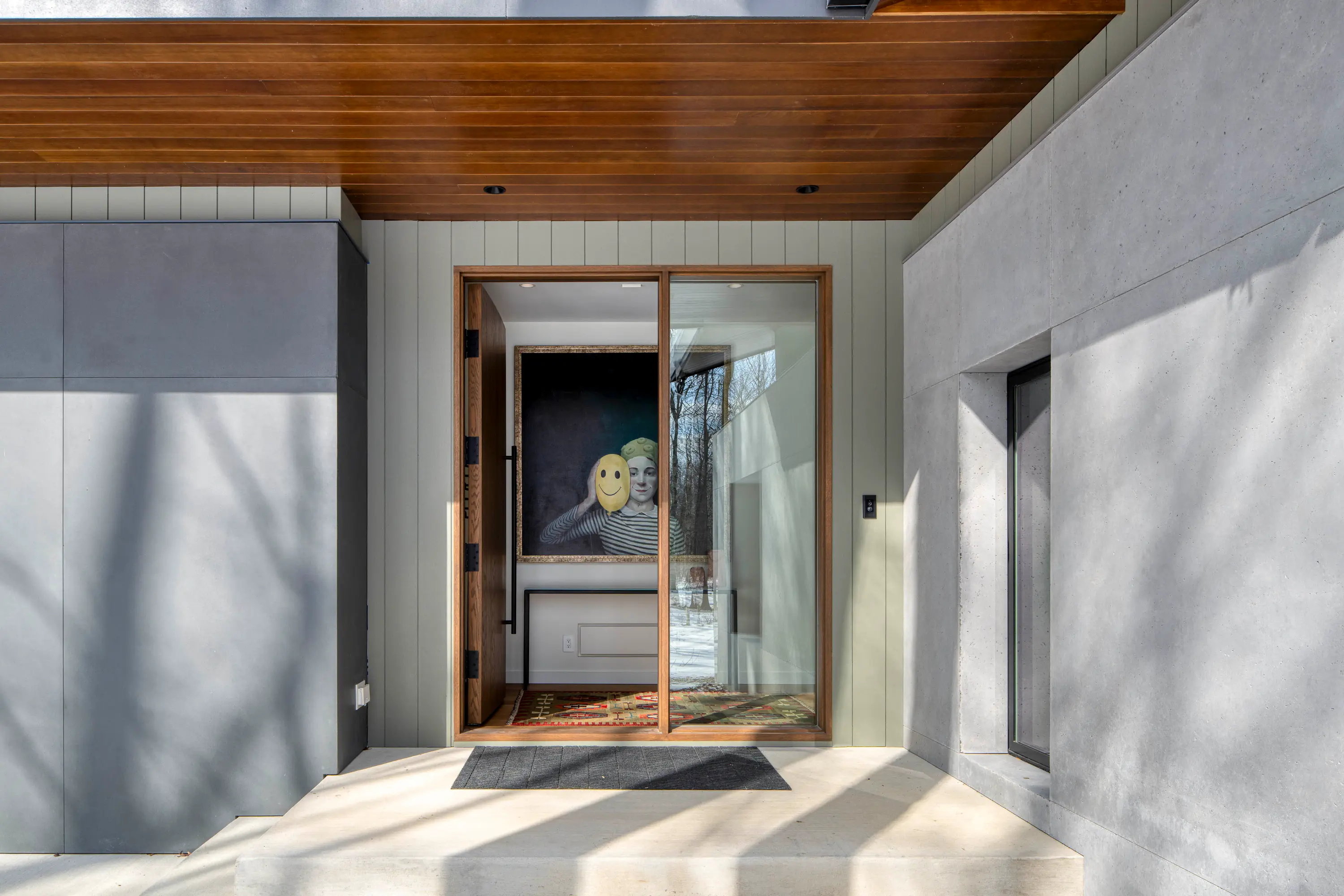 Modern home entry with wooden ceiling, sliding glass door, and a painting of a person holding a smiley face mask.