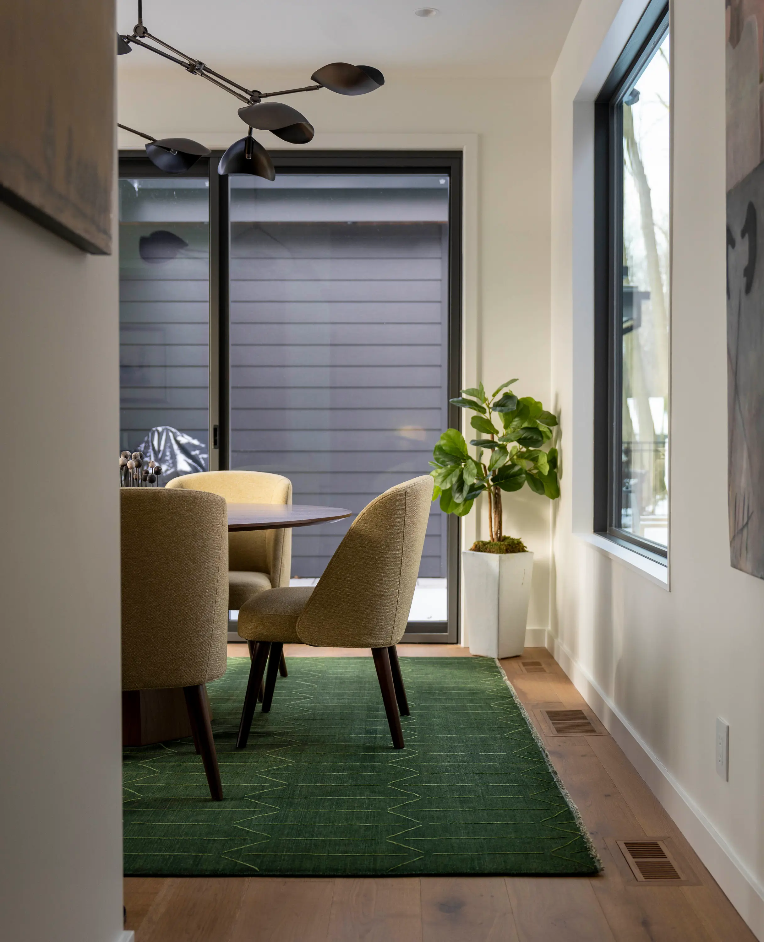 Modern dining area with round table, beige chairs, green rug, large windows, and a potted plant.
