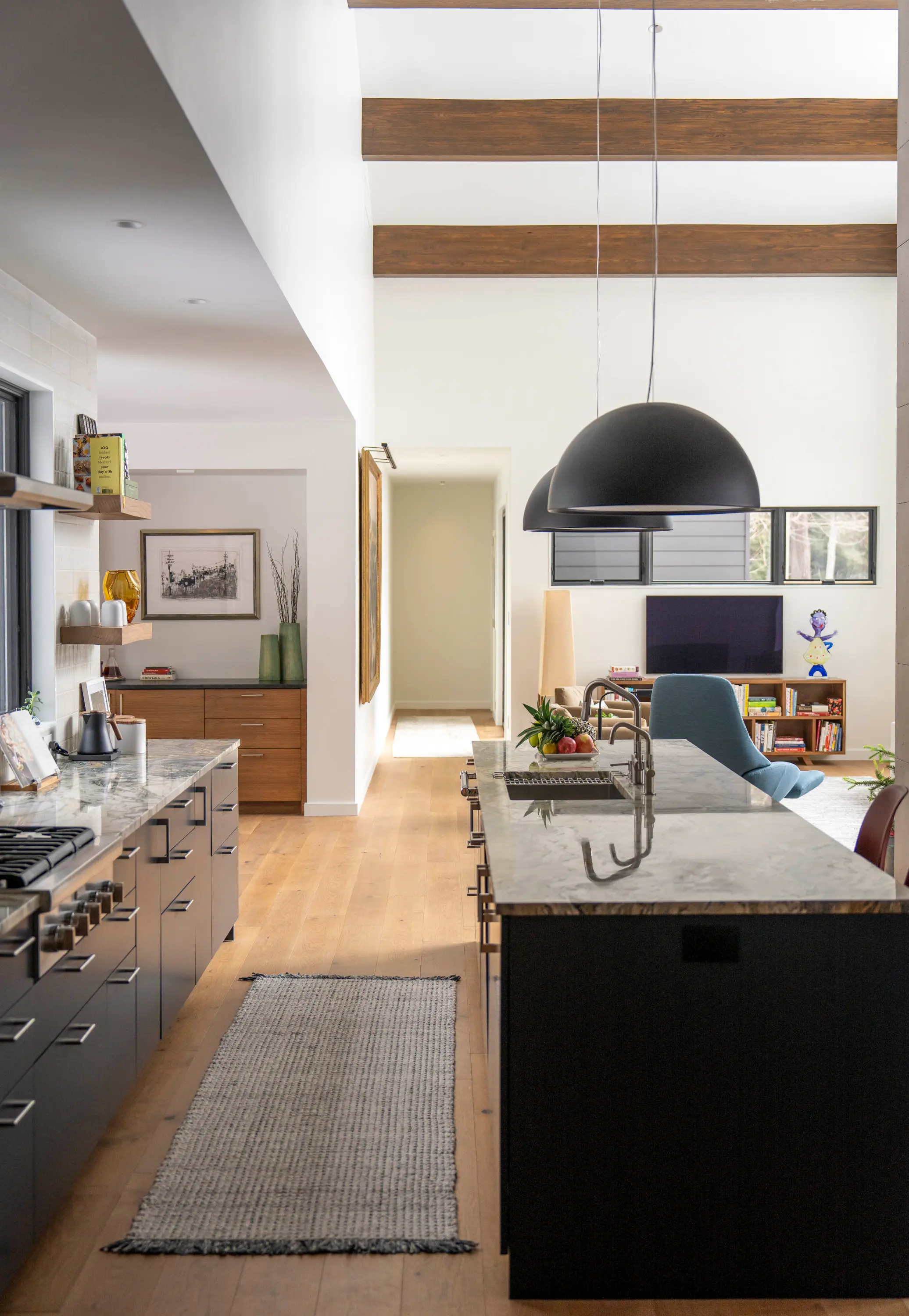 Modern kitchen with marble island, black cabinets, wooden beams, blue chair, and living room in background.