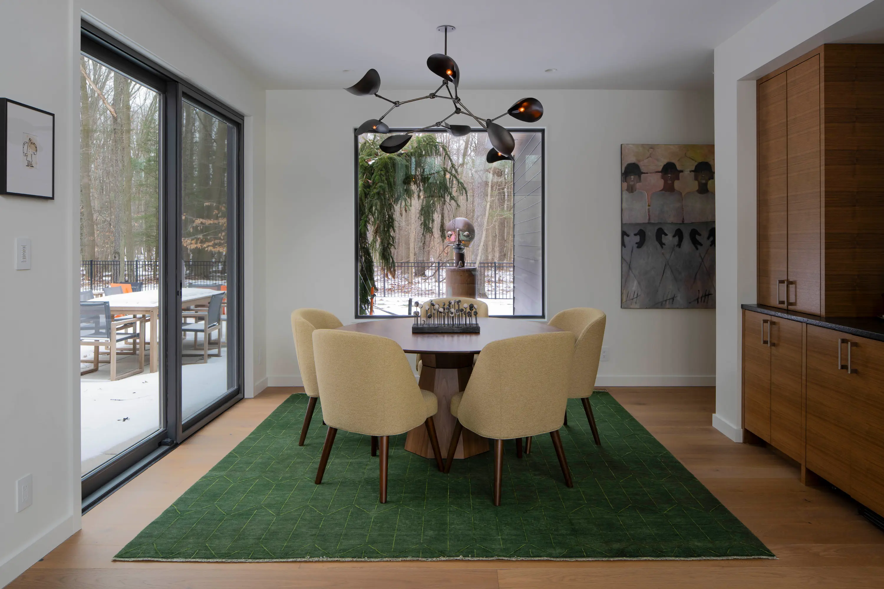 Dining room with round wood table, six beige chairs on green rug, large window, and modern black chandelier.