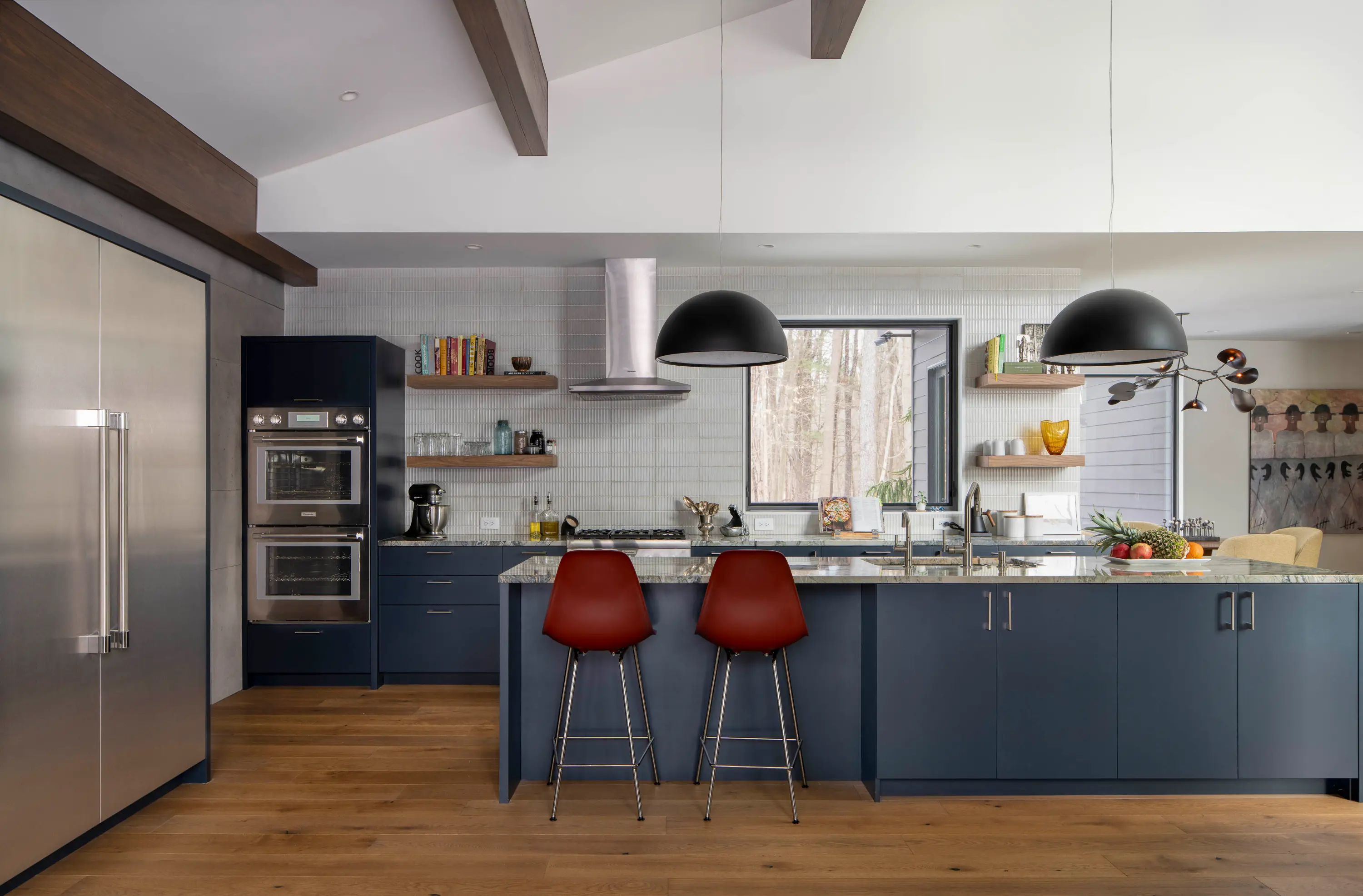 Modern kitchen with blue cabinets, stainless steel appliances, wood floor, red bar stools, and large window.