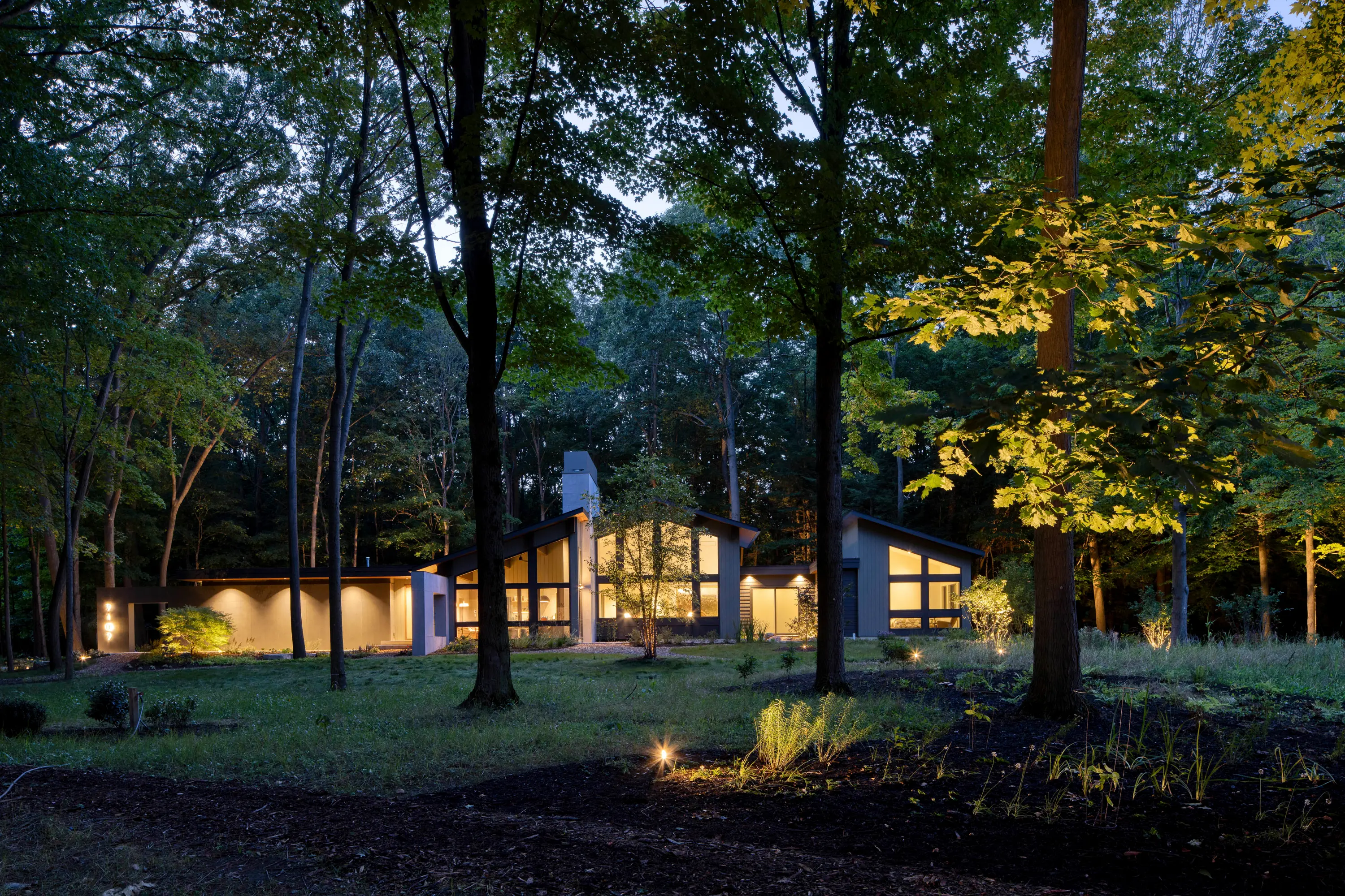 Modern house with large windows lit inside amid tall trees at dusk in a wooded area.