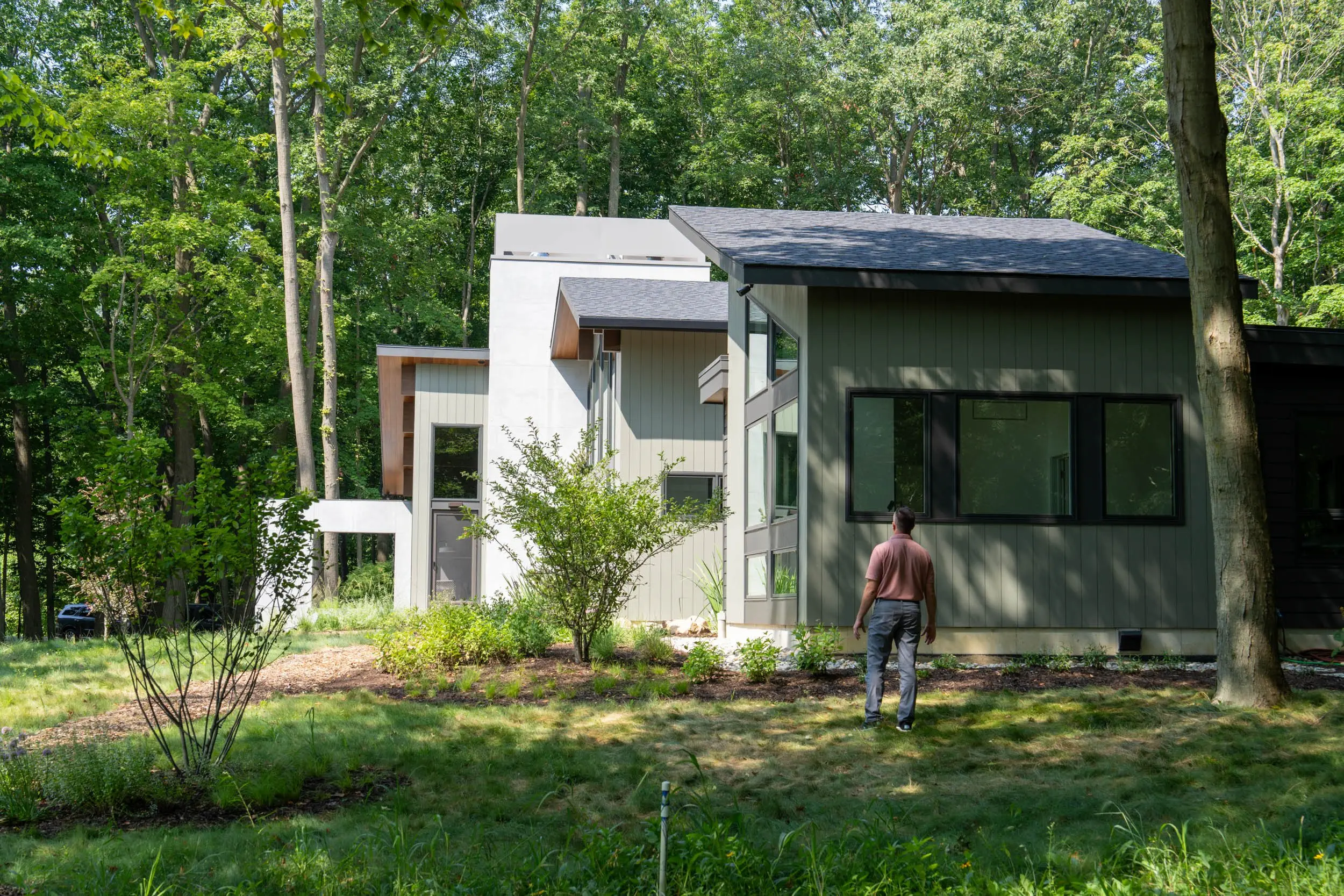 Person in a pink shirt stands on grass viewing a modern house surrounded by trees and plants.