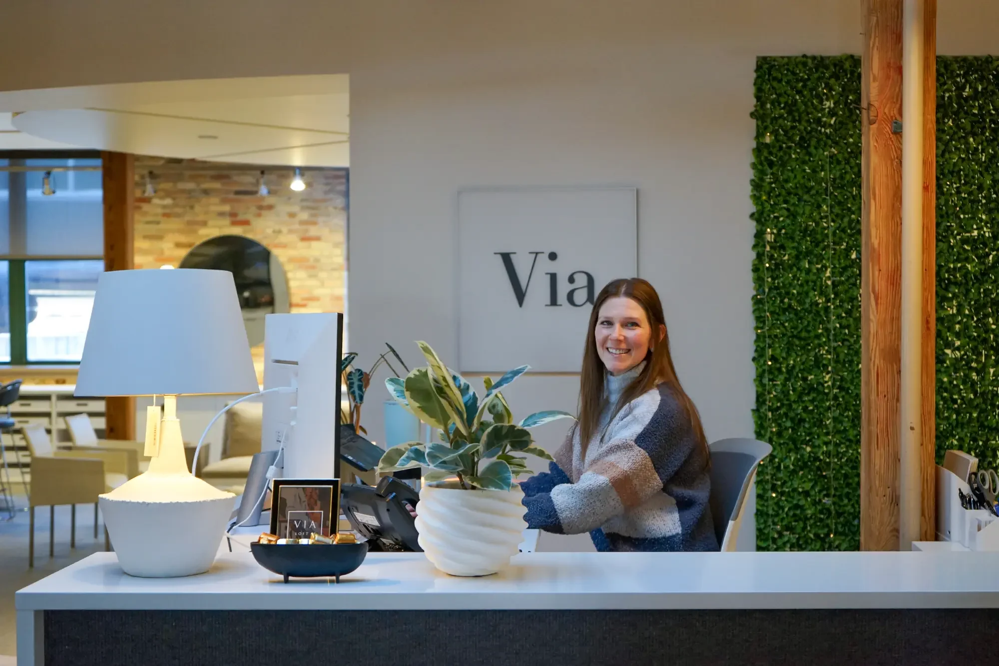 Smiling woman sitting at a reception desk with a plant, lamp, and computer in a modern office.