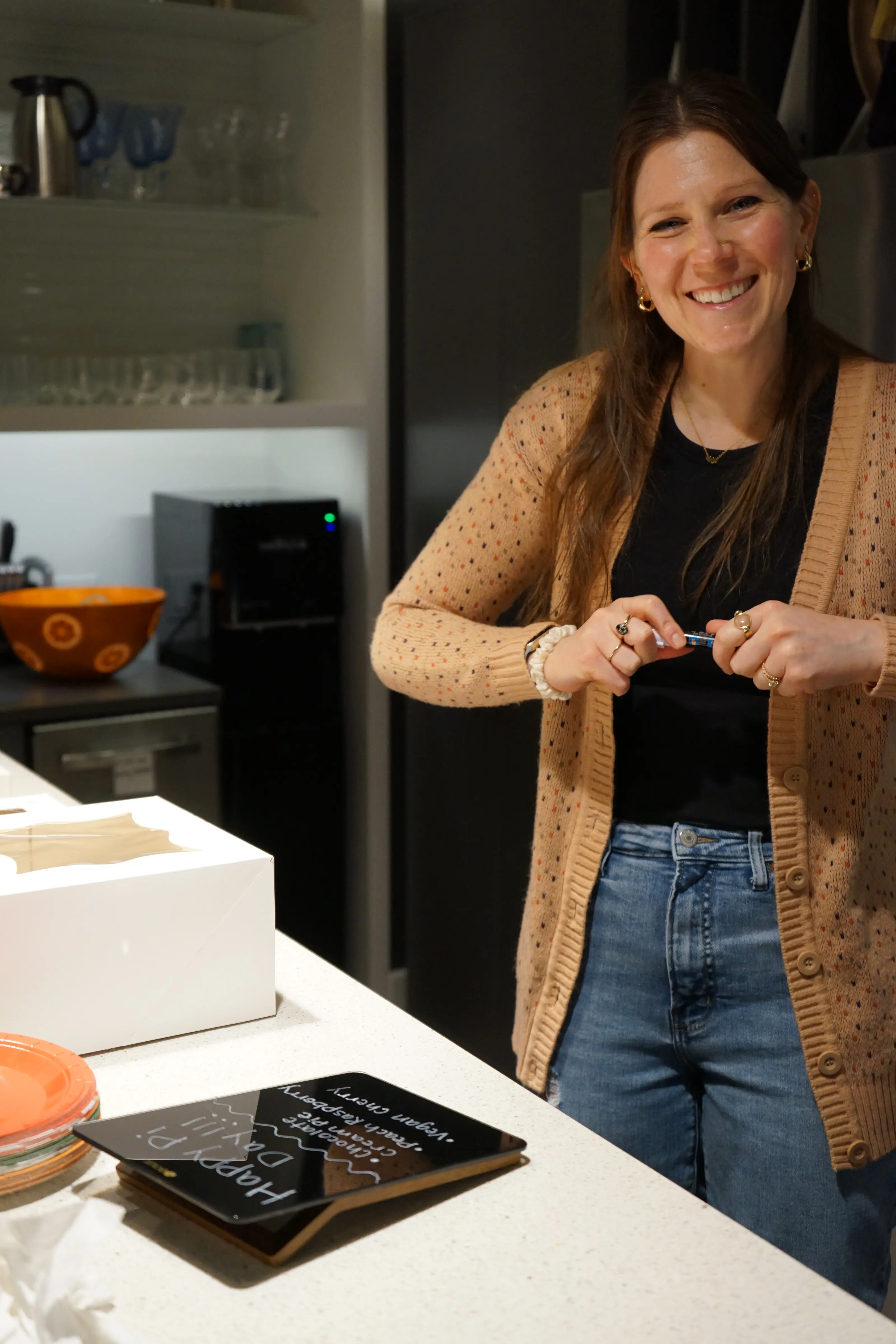 Smiling woman in a brown cardigan and jeans stands in a kitchen near a counter with a menu board.