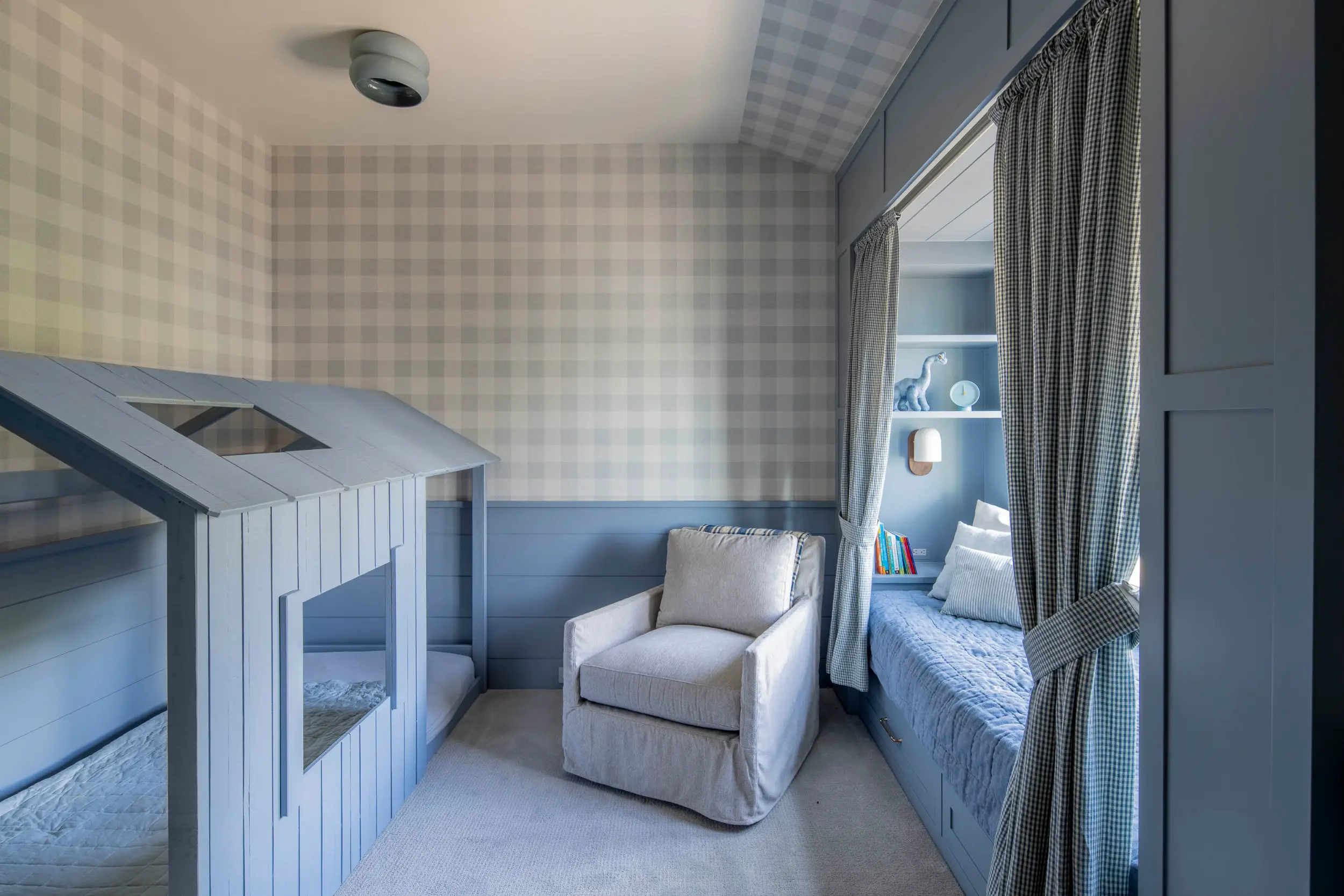 Children's bedroom with blue house-shaped beds, beige armchair, and blue built-in daybed with curtains.