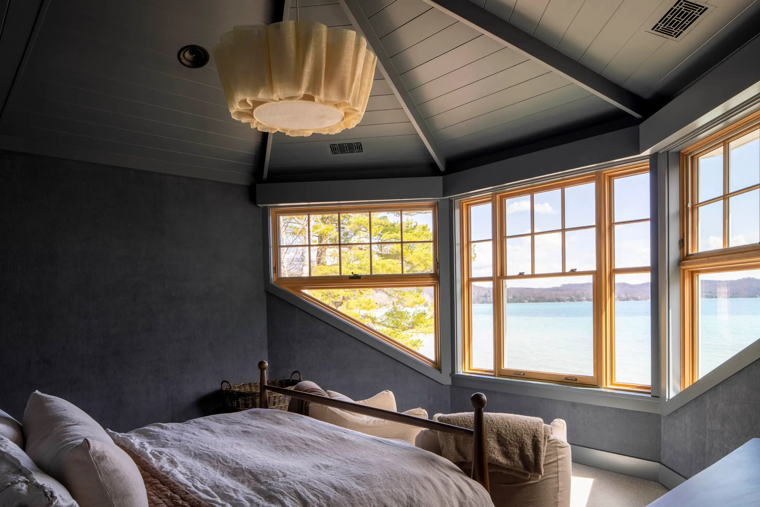 Cozy bedroom with gray walls and ceiling, wooden framed windows overlooking a lake and trees.