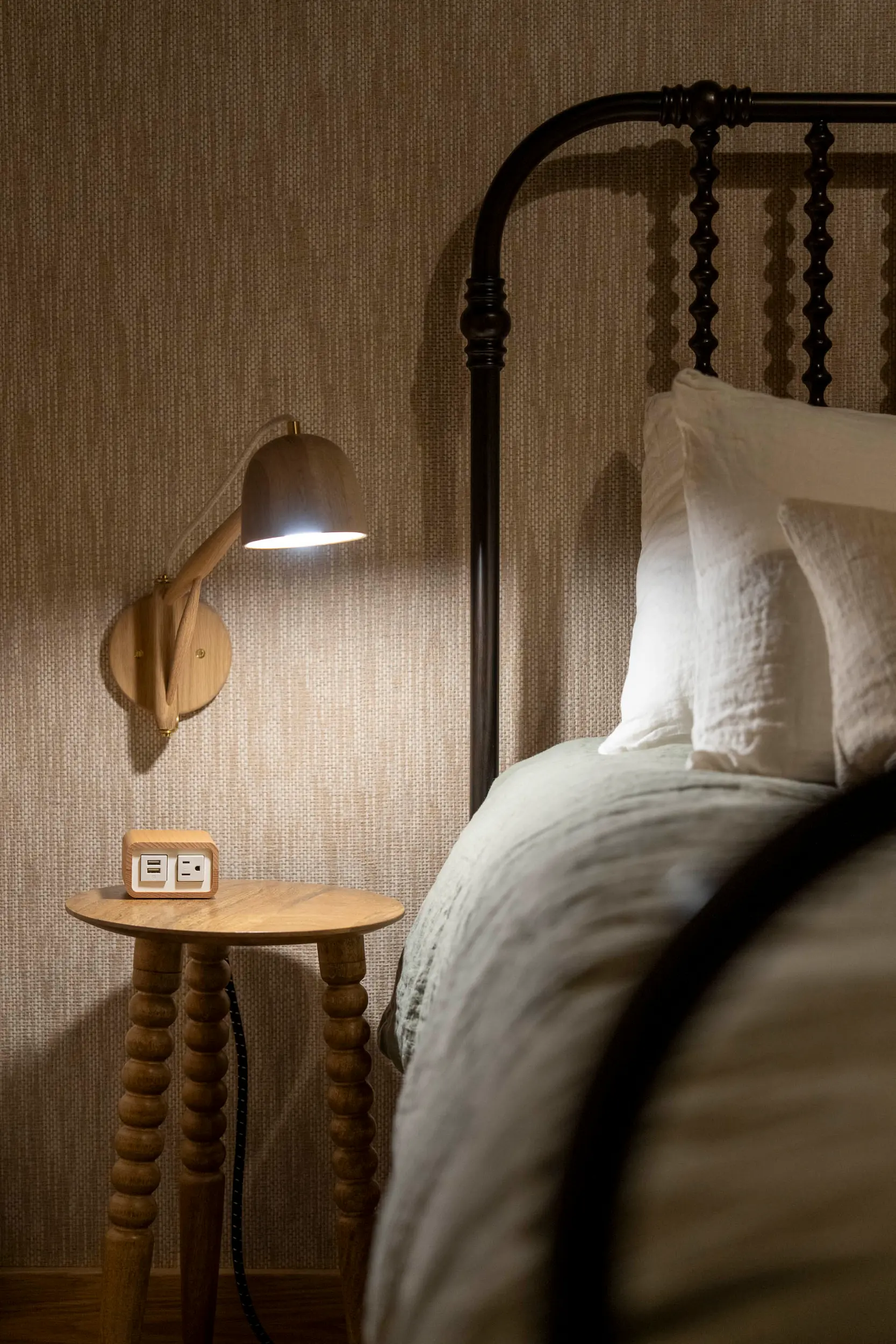 Wooden bedside table with power outlet and lit wooden wall lamp next to a bed with white pillows.