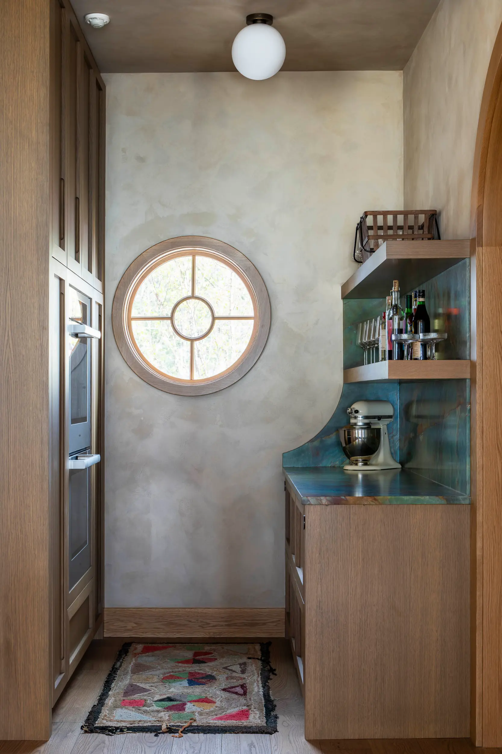 Small kitchen area with wood cabinets, a round window, patterned rug, mixer, and shelves with bottles and glasses.