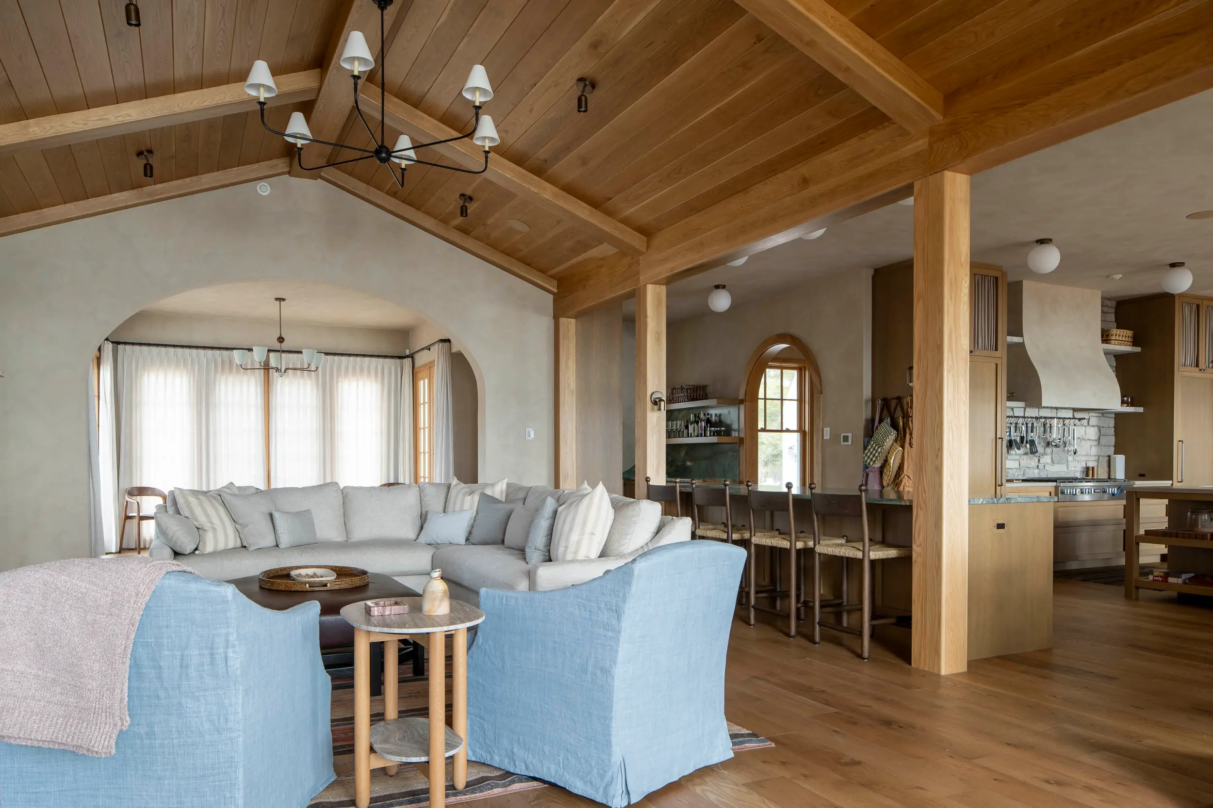 Cozy living room with light gray sectional sofa, blue armchairs, wooden floors, and an open kitchen in the back.