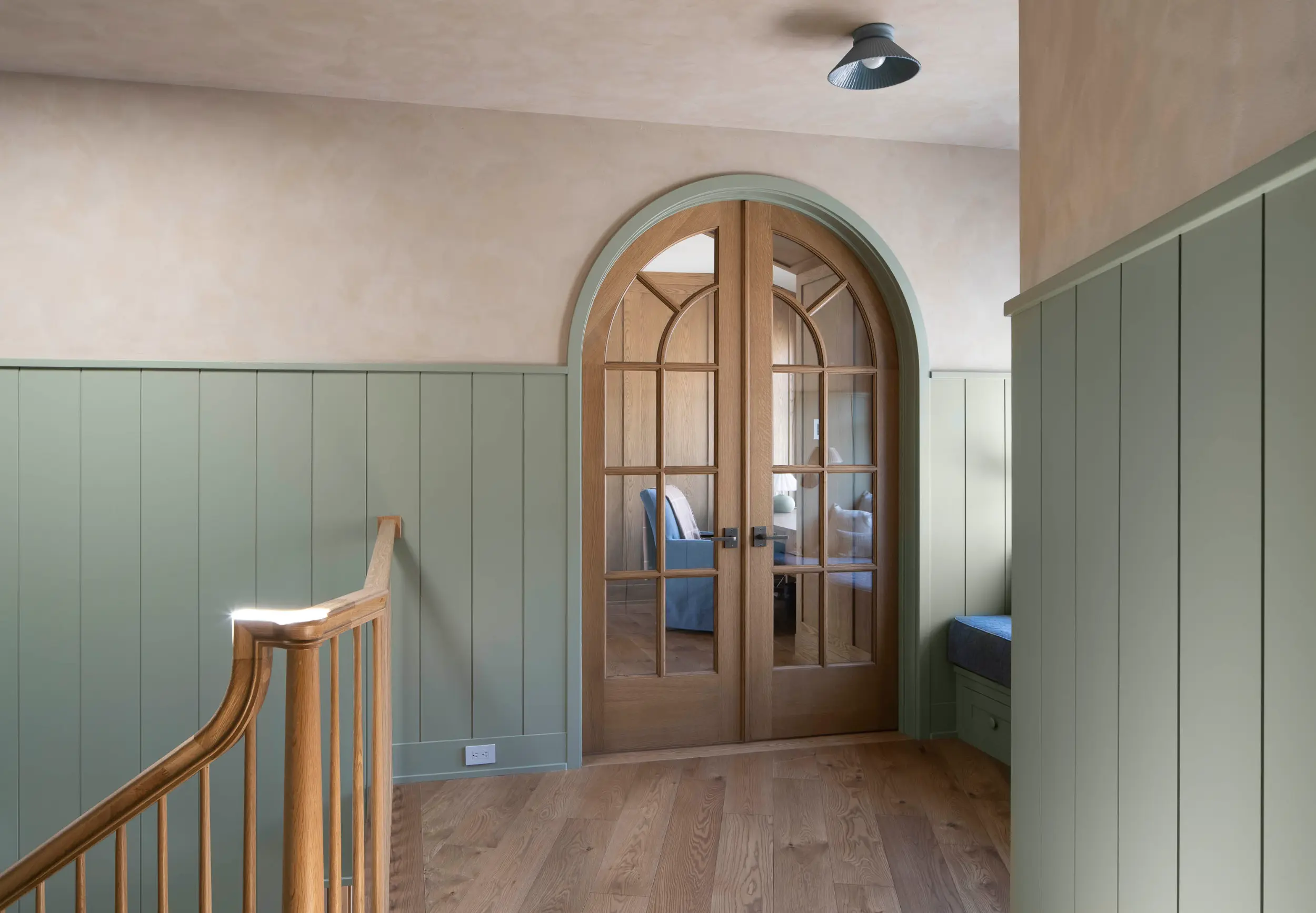 Hallway with green paneled walls, wooden stairs, and arched double glass doors revealing a room with chairs.