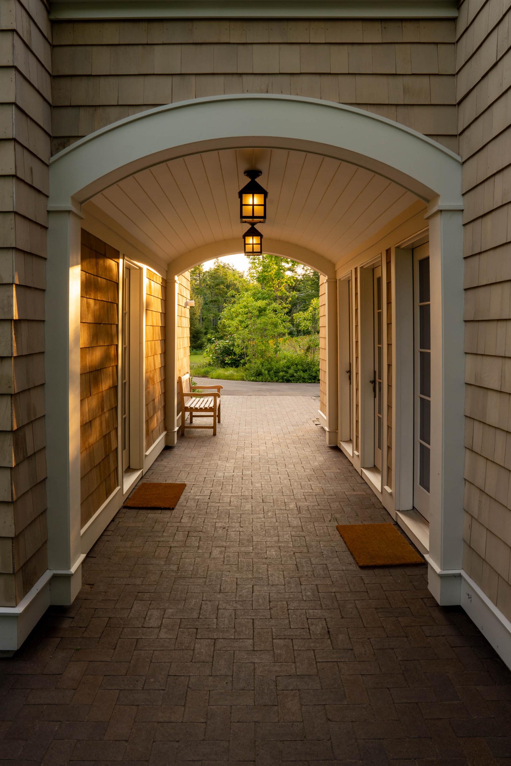 Covered walkway with two lanterns, wooden bench, and doors leading outside to greenery at sunset.