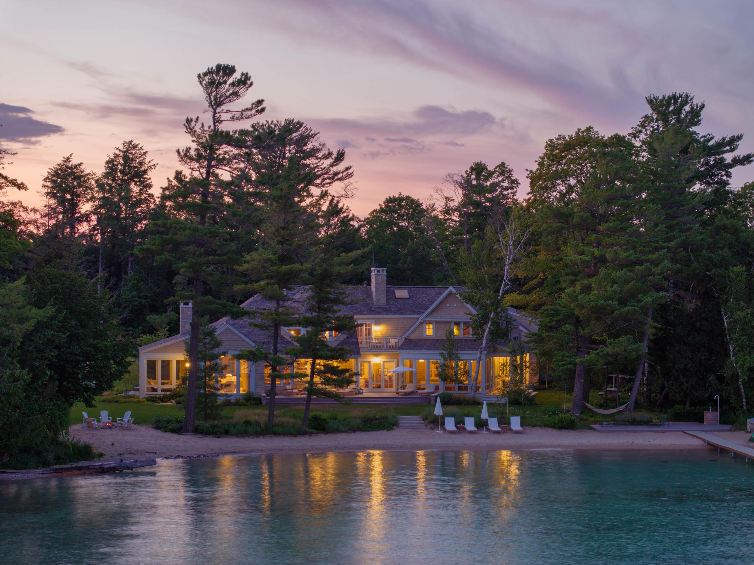 Lakefront house at sunset with lit windows, surrounded by trees and beach chairs near clear water.
