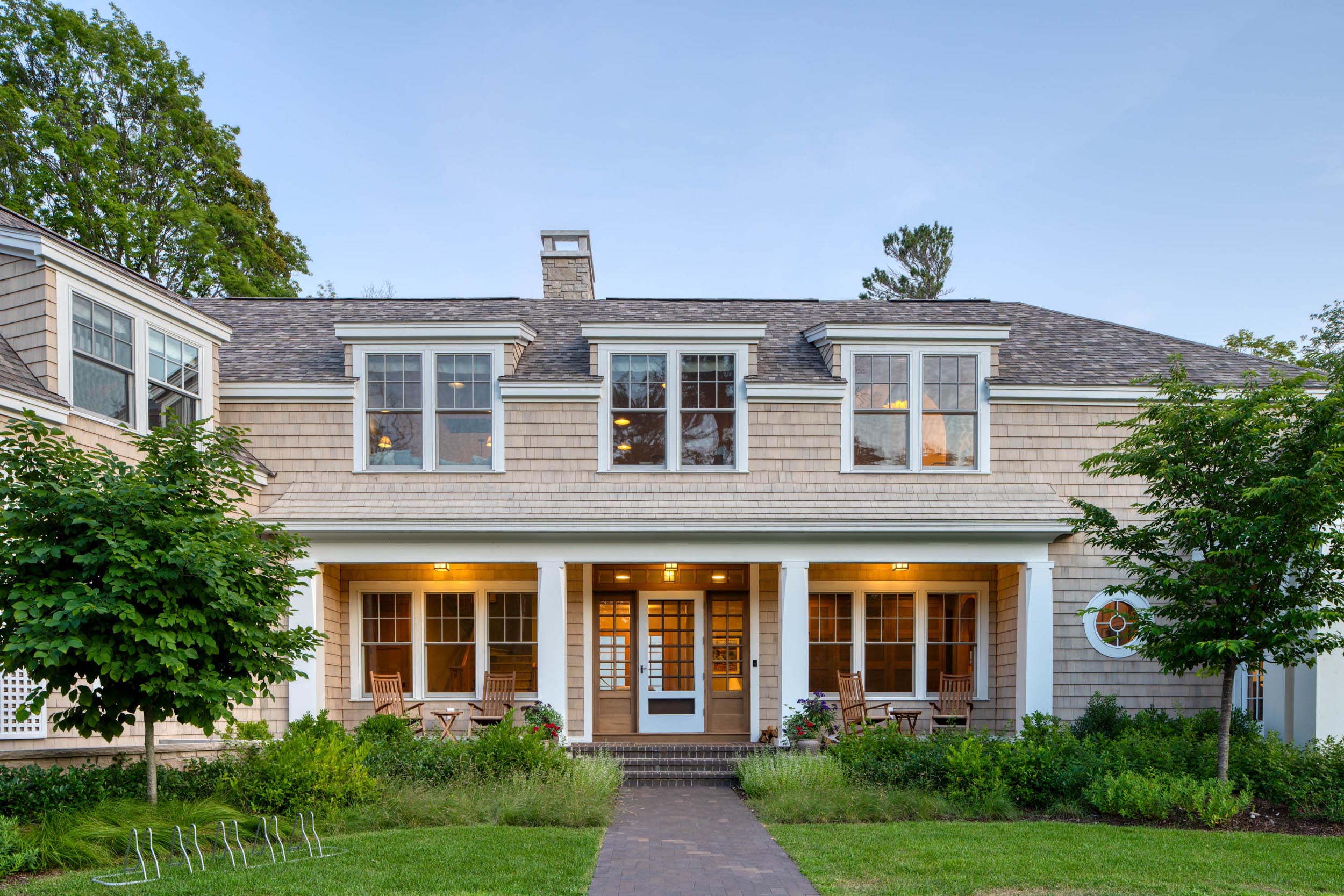Front view of a two-story house with beige siding, white trim, a porch, chairs, and green lawn.