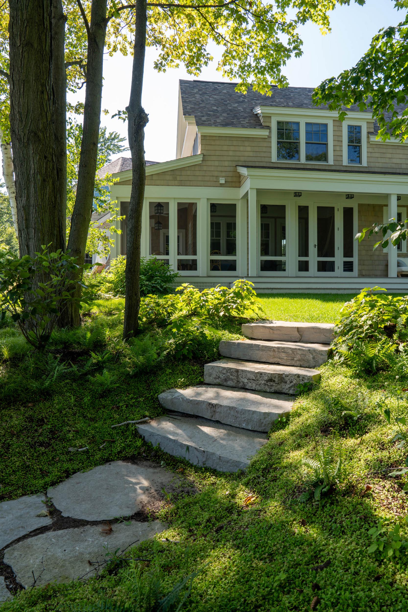 Stone steps in lush green garden leading up to a screened porch on a wood-shingle house.