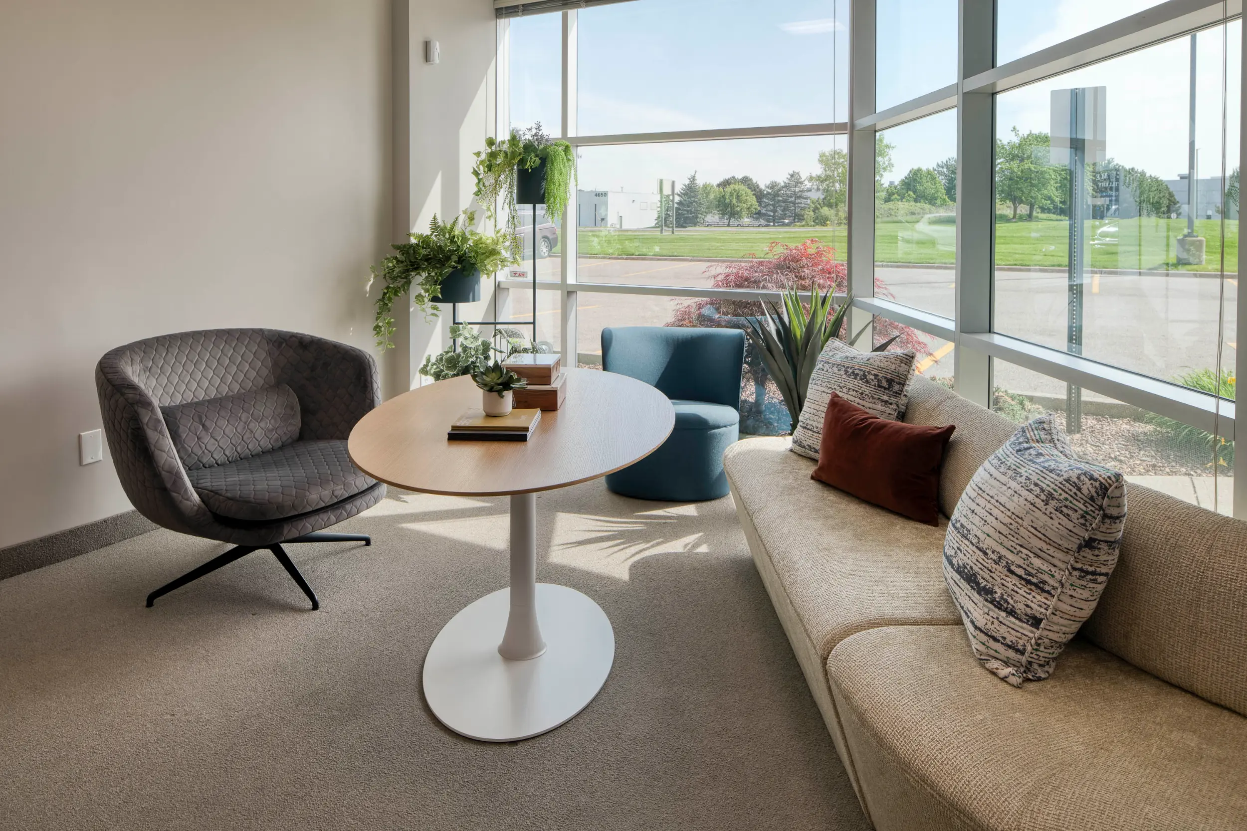 Modern lounge with a beige sofa, patterned pillows, round table, gray armchair, and large windows.
