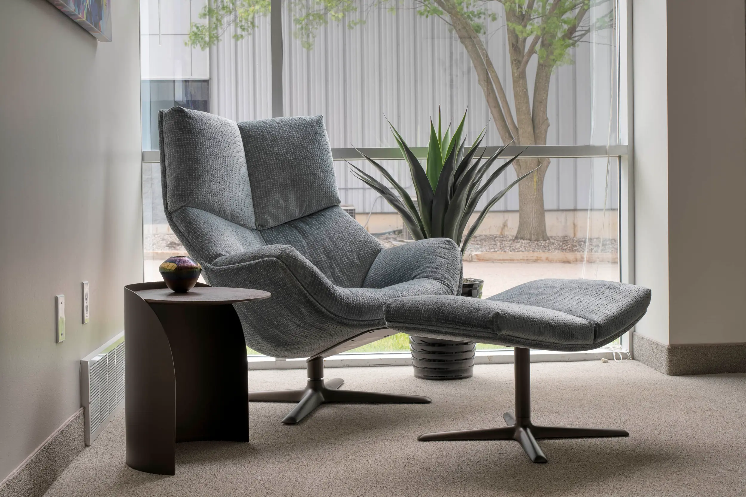 Gray cushioned lounge chair with matching ottoman next to a side table and a potted plant by a window.