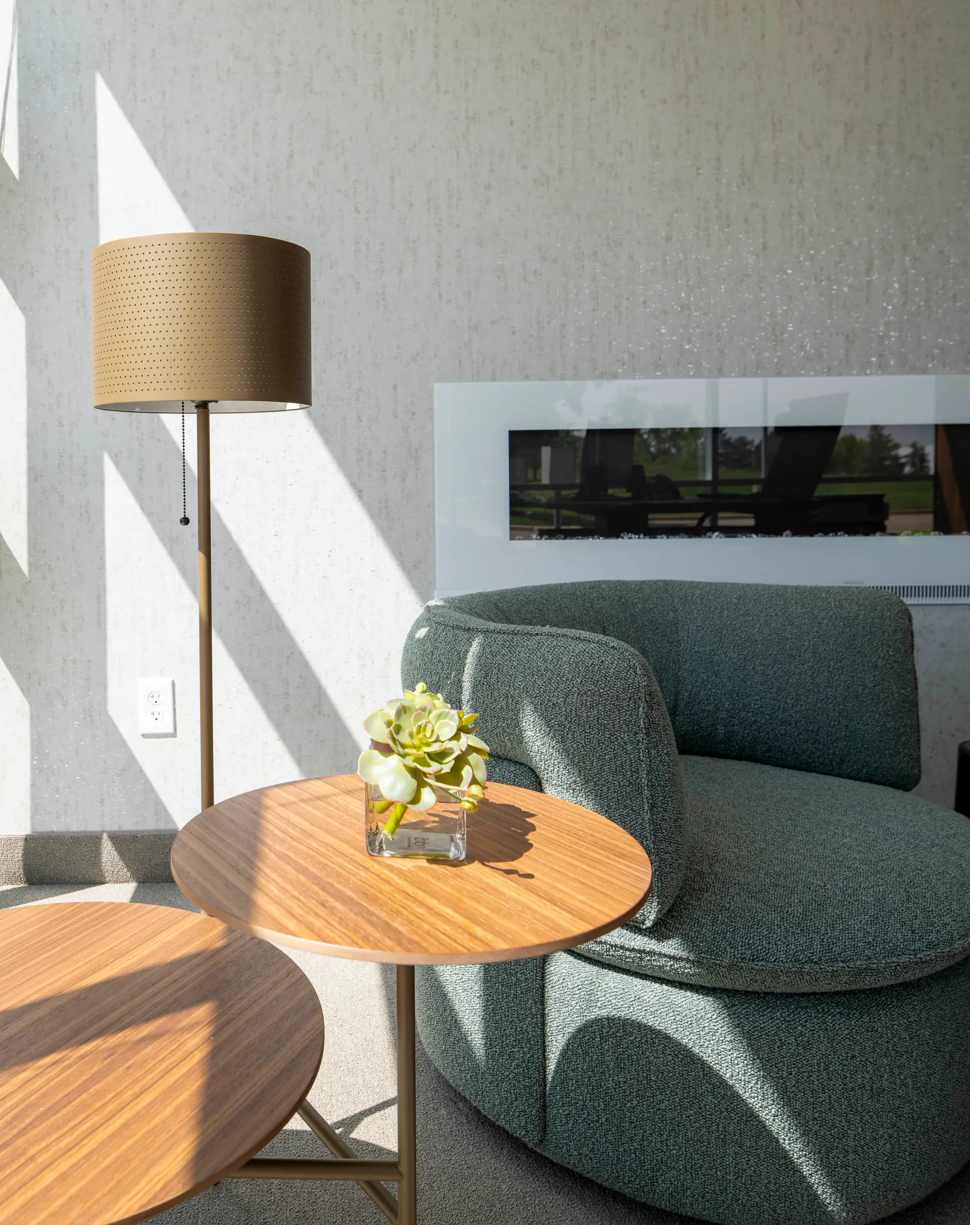 Modern living room corner with curved green armchair, two round wooden tables, lamp, and small plant.