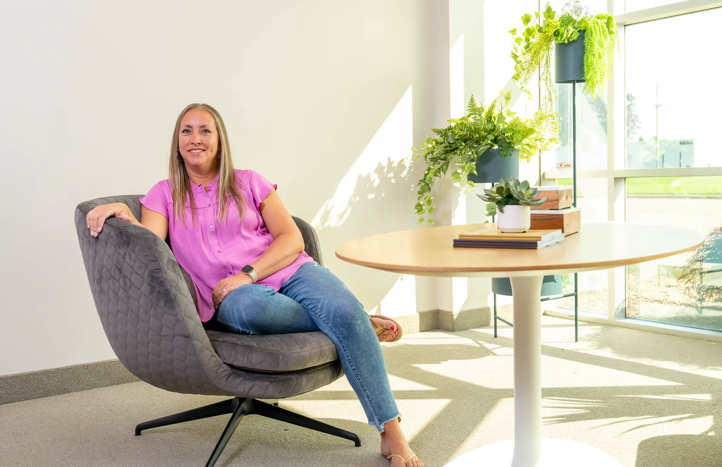 Woman in pink shirt and jeans sits in gray chair next to round table with green plants by window.