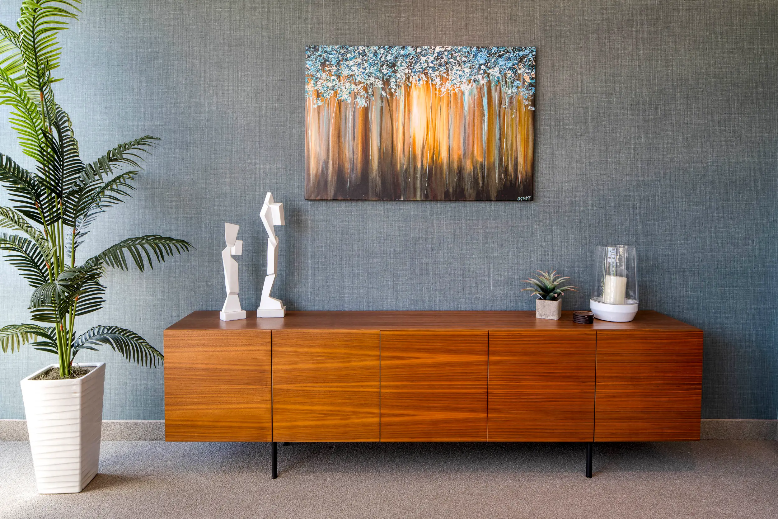 Wooden sideboard against blue wall with abstract art, white sculptures, plant, and candle decor.