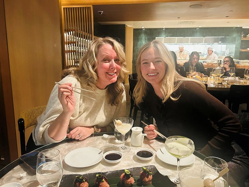 Two women smiling with chopsticks at a table with sushi, drinks, and soy sauce dishes in a restaurant.