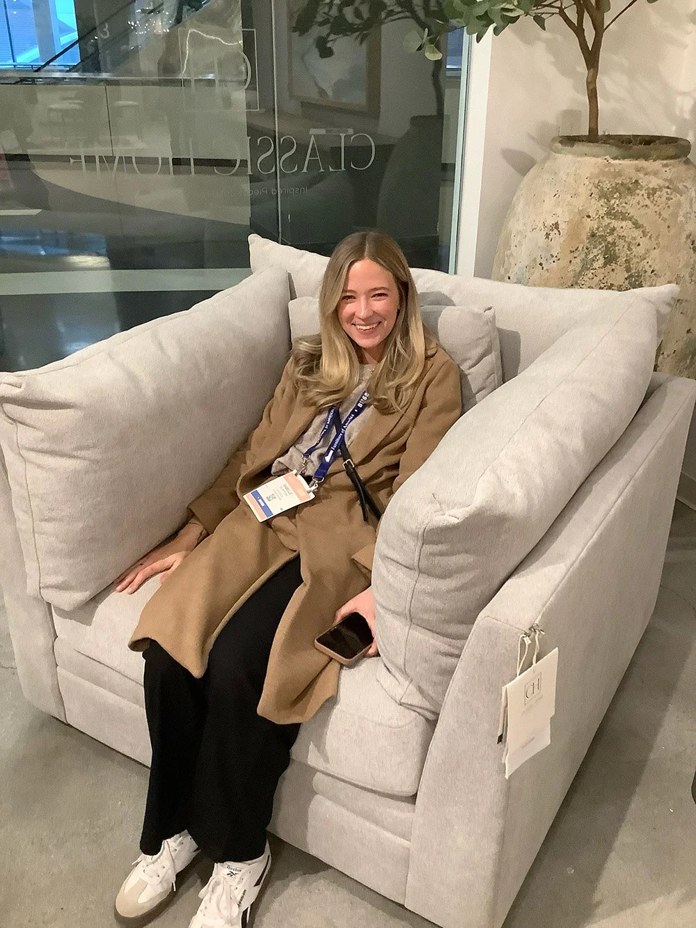 Smiling woman in a brown coat sitting comfortably on a gray cushioned sofa holding a phone.