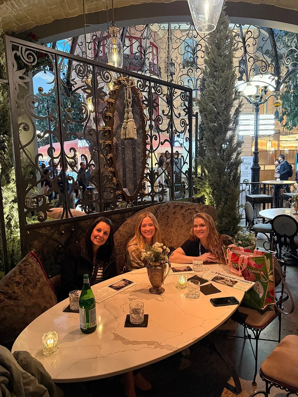 Three women sitting at a marble table in a cozy, ornate outdoor cafe with candles and menus.