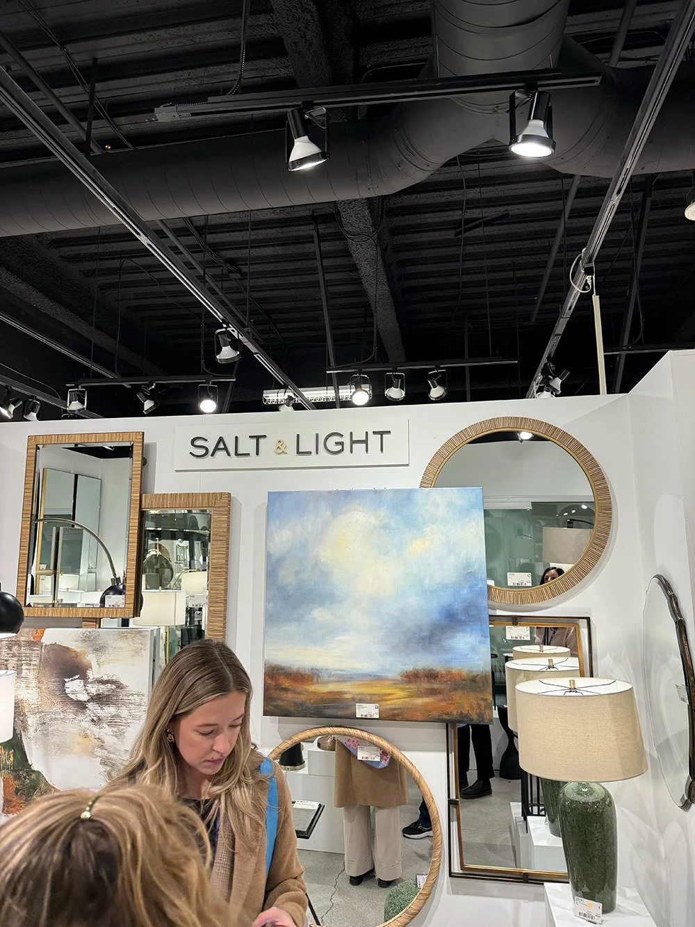 Interior of a showroom with mirrors, a landscape painting, a green lamp, and people browsing.