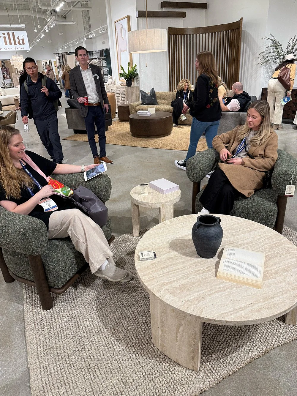 Furniture store scene with two women seated on armchairs and several people walking around displays.