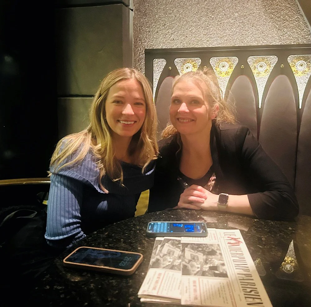 Two women smiling seated at a table with phones and some printed papers in a dimly lit restaurant.