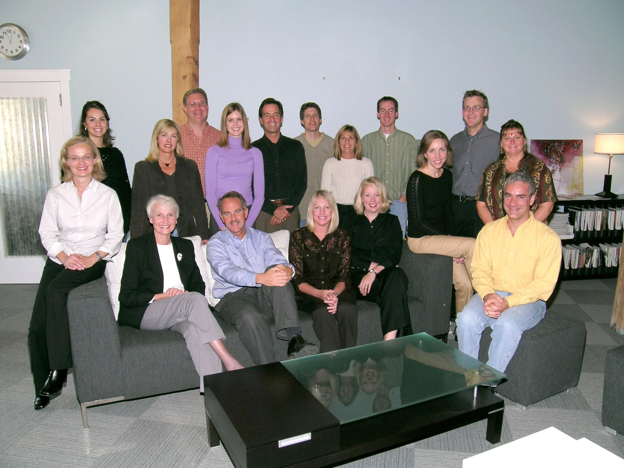 Group of 16 adults posing indoors on couches and chairs, smiling at the camera.