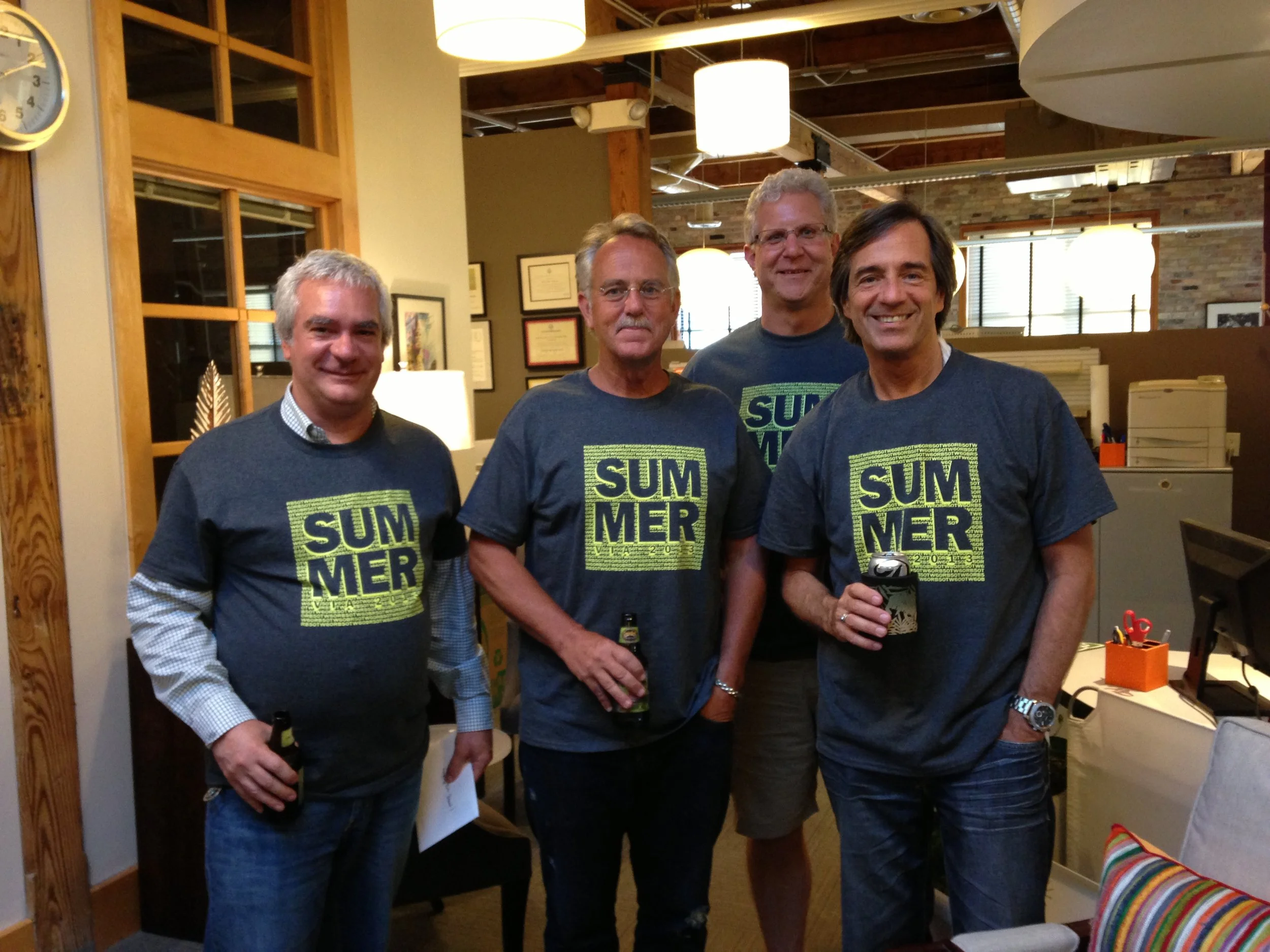 Four men smiling indoors wearing matching 'SUMMER' t-shirts and holding drinks, posing for a photo.