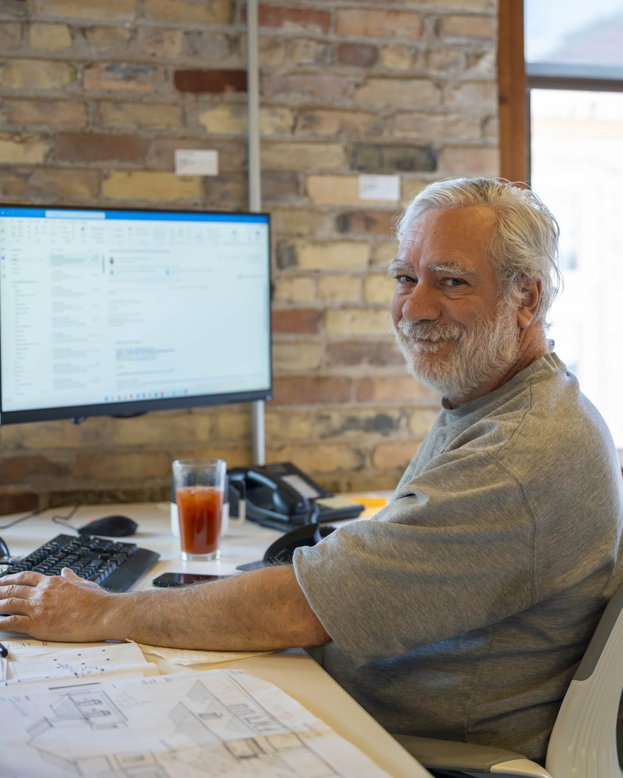 Older man with white hair and beard smiles at camera while working at a desk with blueprints and computer.
