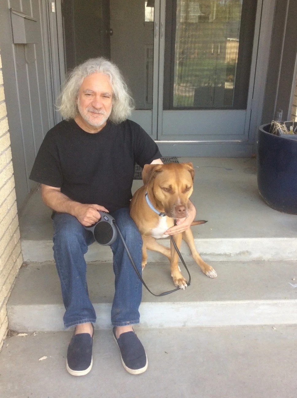 Man with gray hair in black shirt sitting on steps next to a brown dog wearing a blue collar.