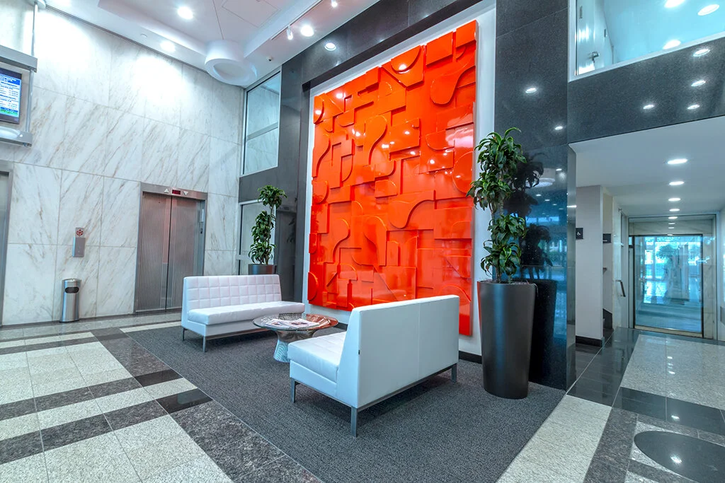 Modern lobby with white seating, gray carpet, large orange abstract wall art, and potted plants.