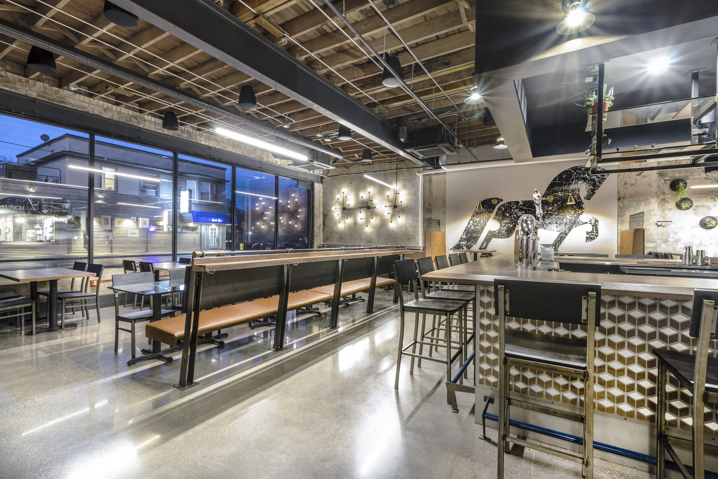 Modern empty bar with long wooden tables, high chairs, decorative lights, and a horse mural on the wall.