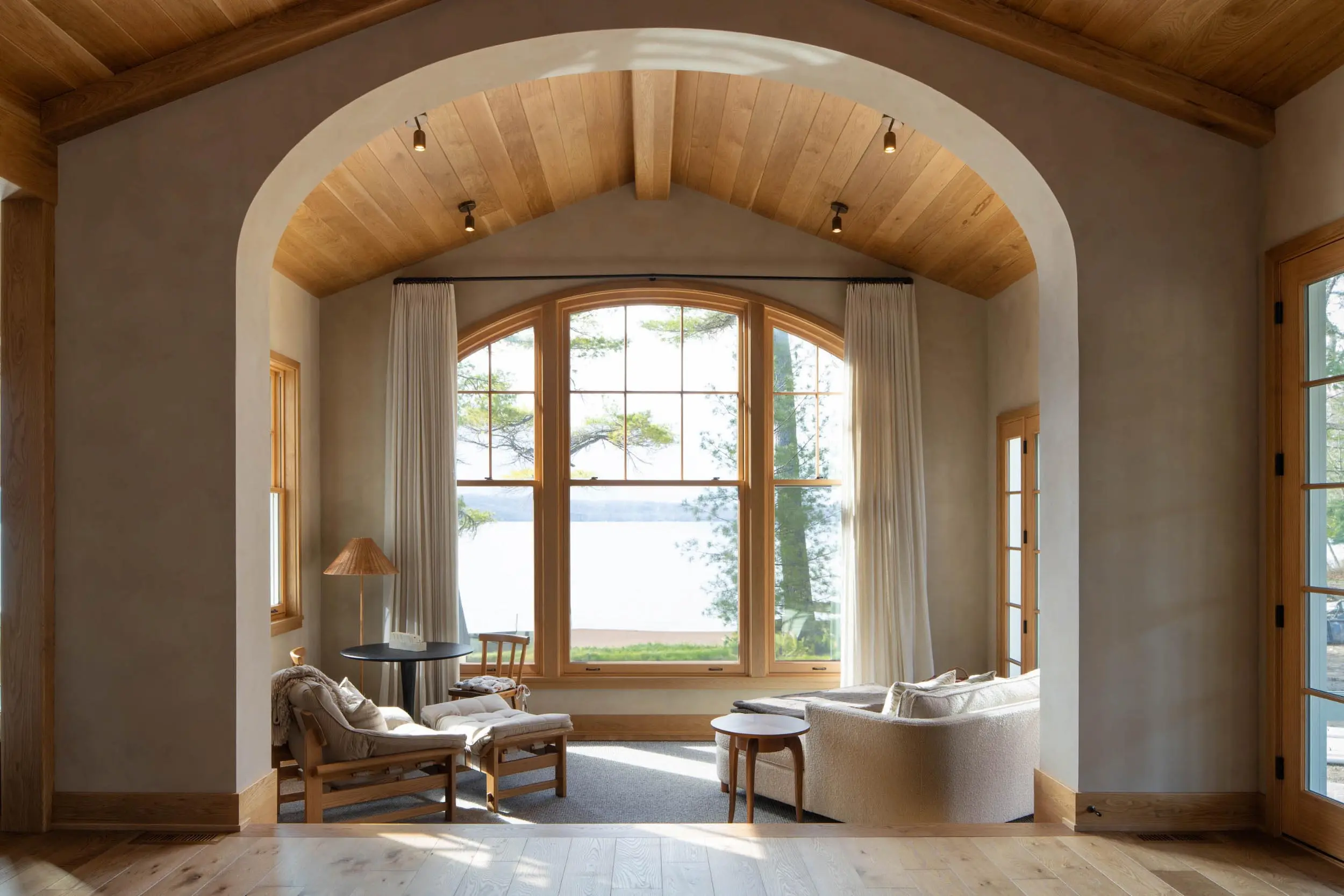 Cozy living room with large arched window overlooking trees and lake, featuring soft sofas and wooden furniture.