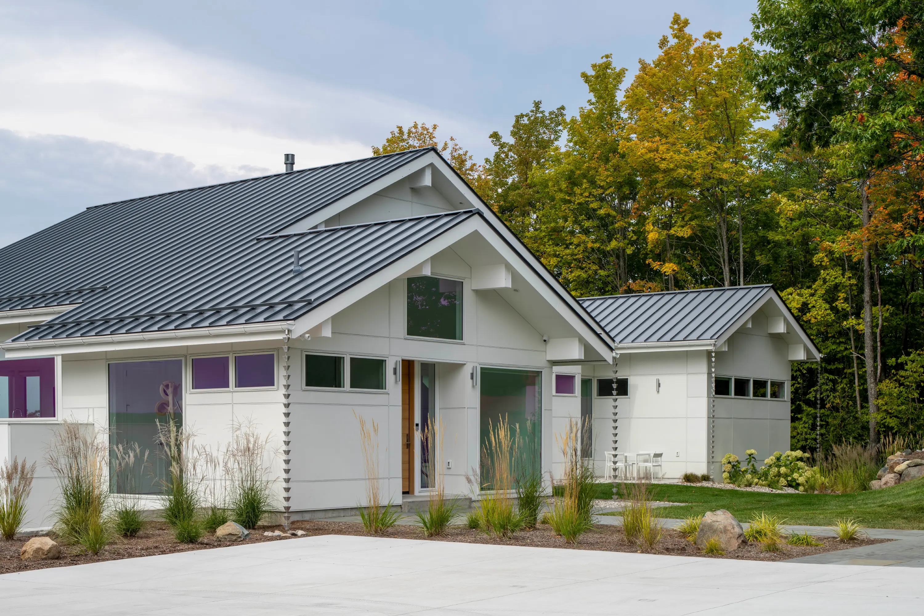 Modern white Scandinavian cottage with metal roof surrounded by fall trees and landscaped yard.