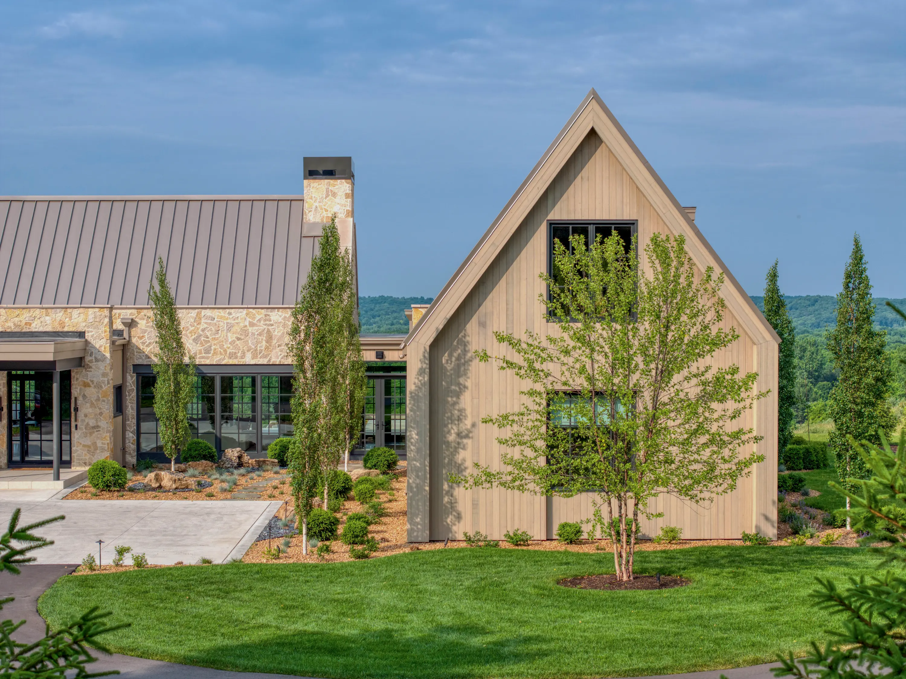 Modern house with stone and wooden exterior, surrounded by green lawn and young trees.