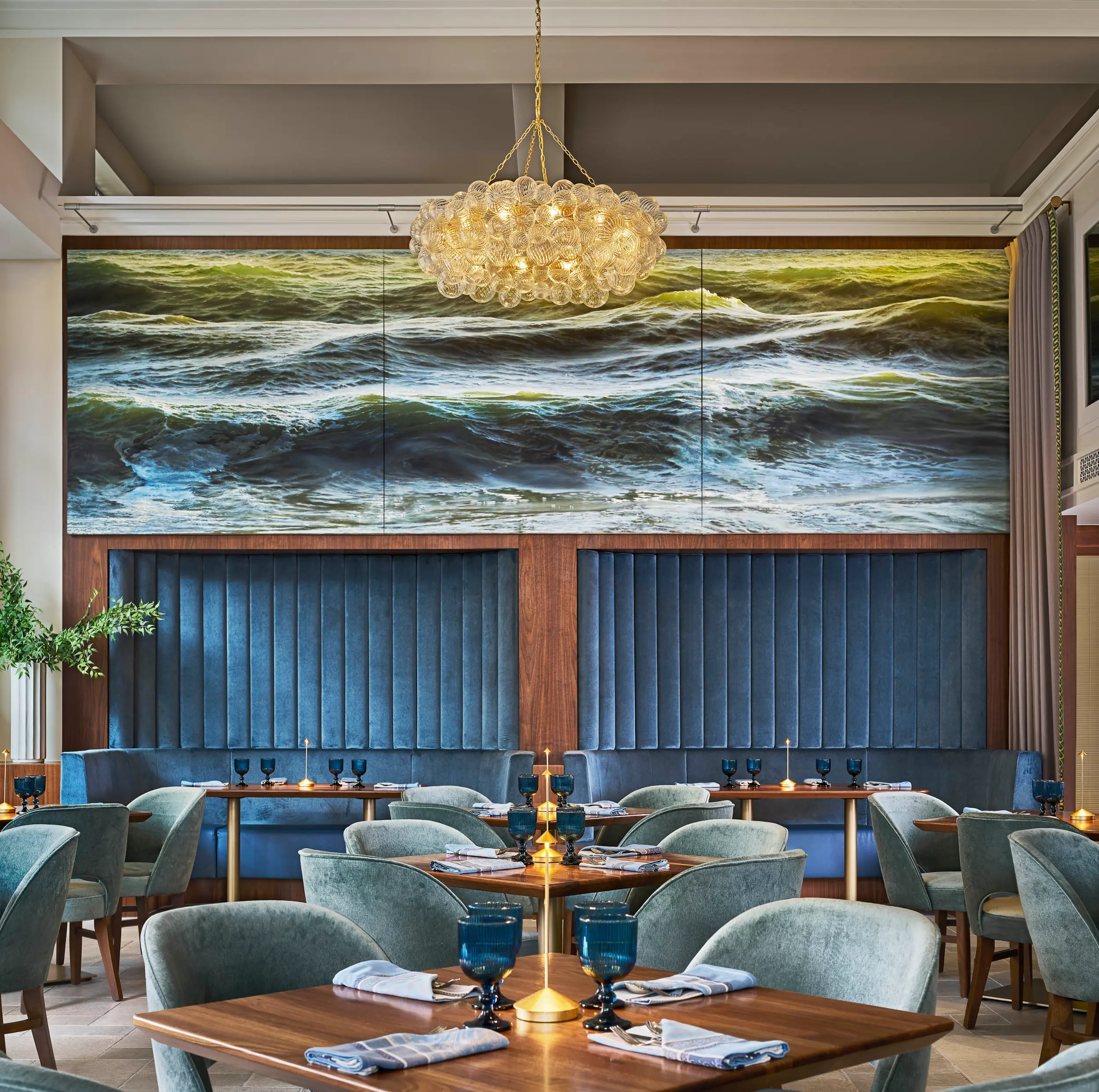 Elegant restaurant interior with blue velvet chairs and wave artwork on the wall under a glass chandelier.