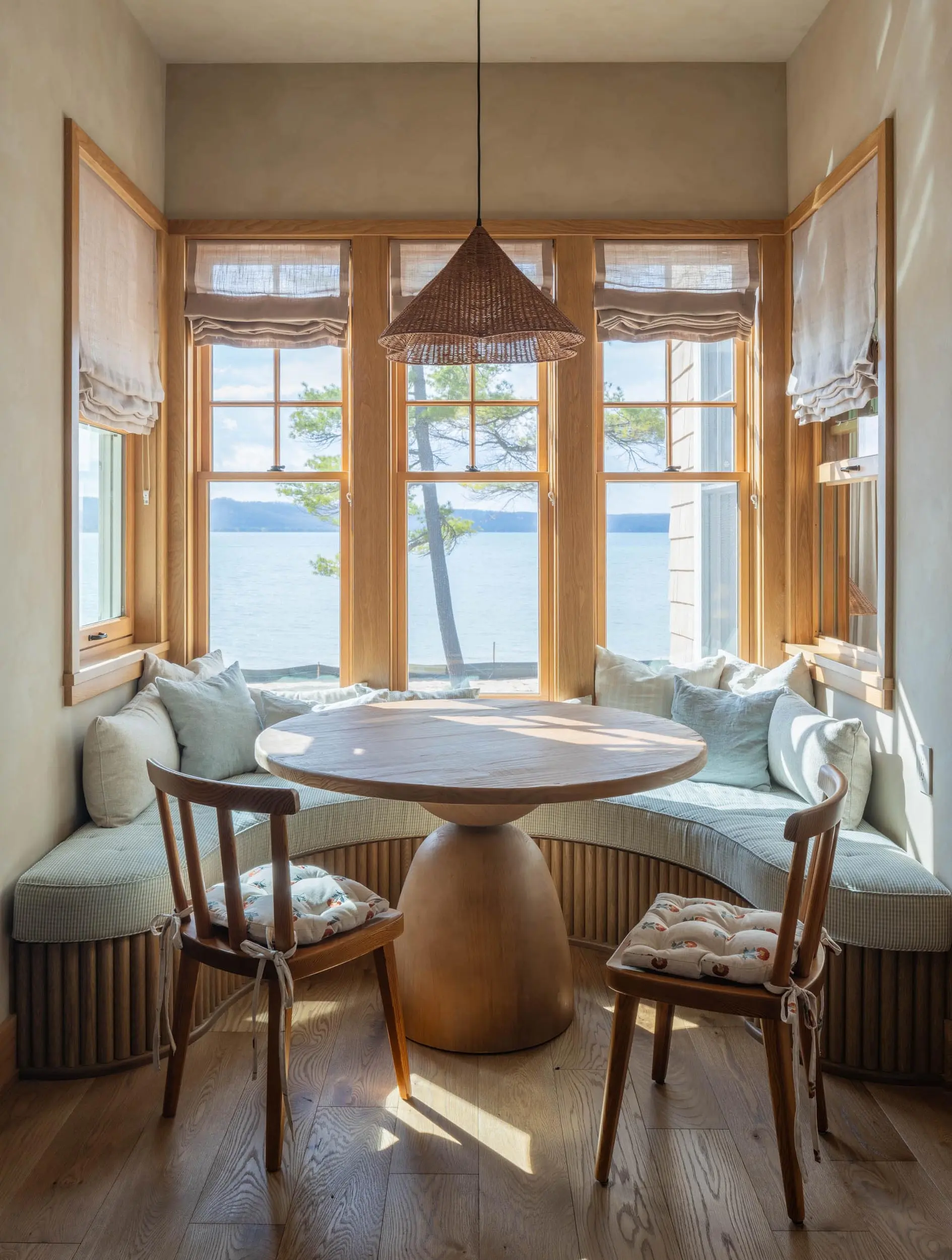 Cozy breakfast nook with round wooden table, cushioned chairs, bench seating, and windows overlooking water.