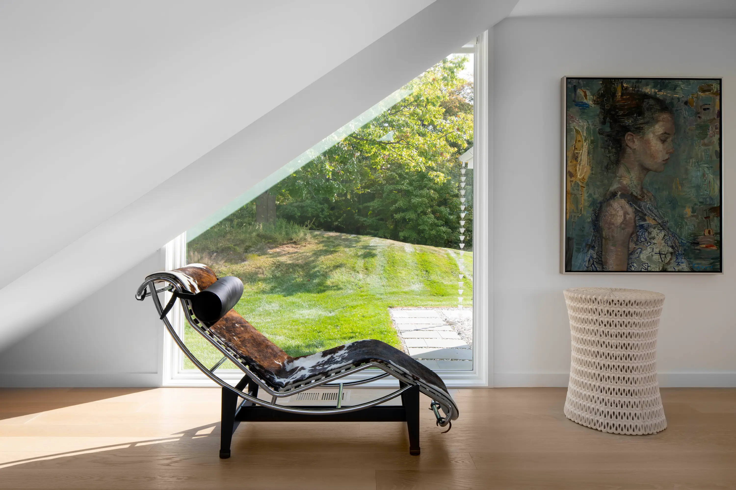 Modern lounge chair with cowhide upholstery beside large window showing green lawn and trees.