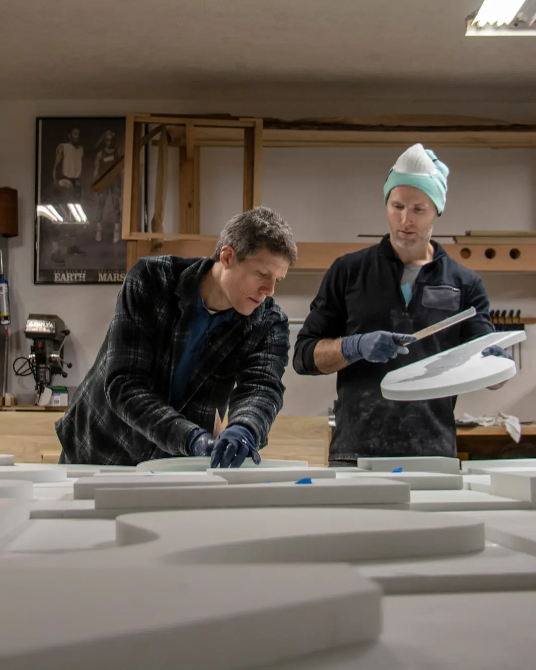 Two men working in a workshop handling round and rectangular white foam pieces on a table.
