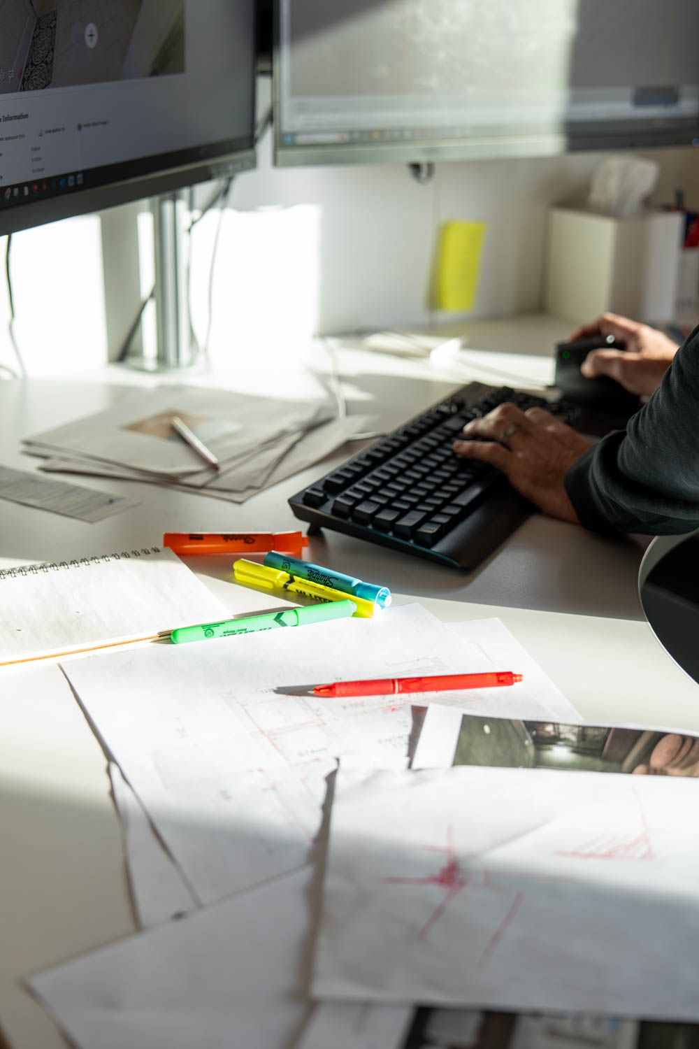 Hands typing on keyboard beside scattered papers, pens, and dual monitors on a bright desk.