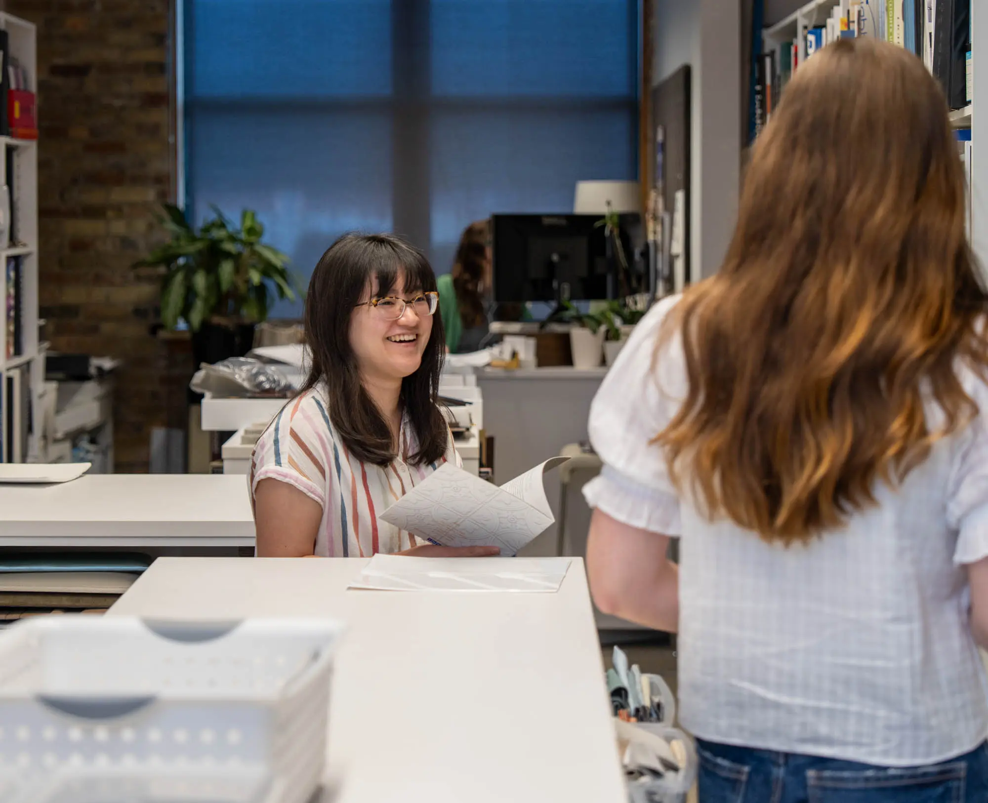 Two women in a modern office; one smiling with papers, the other standing with long hair.
