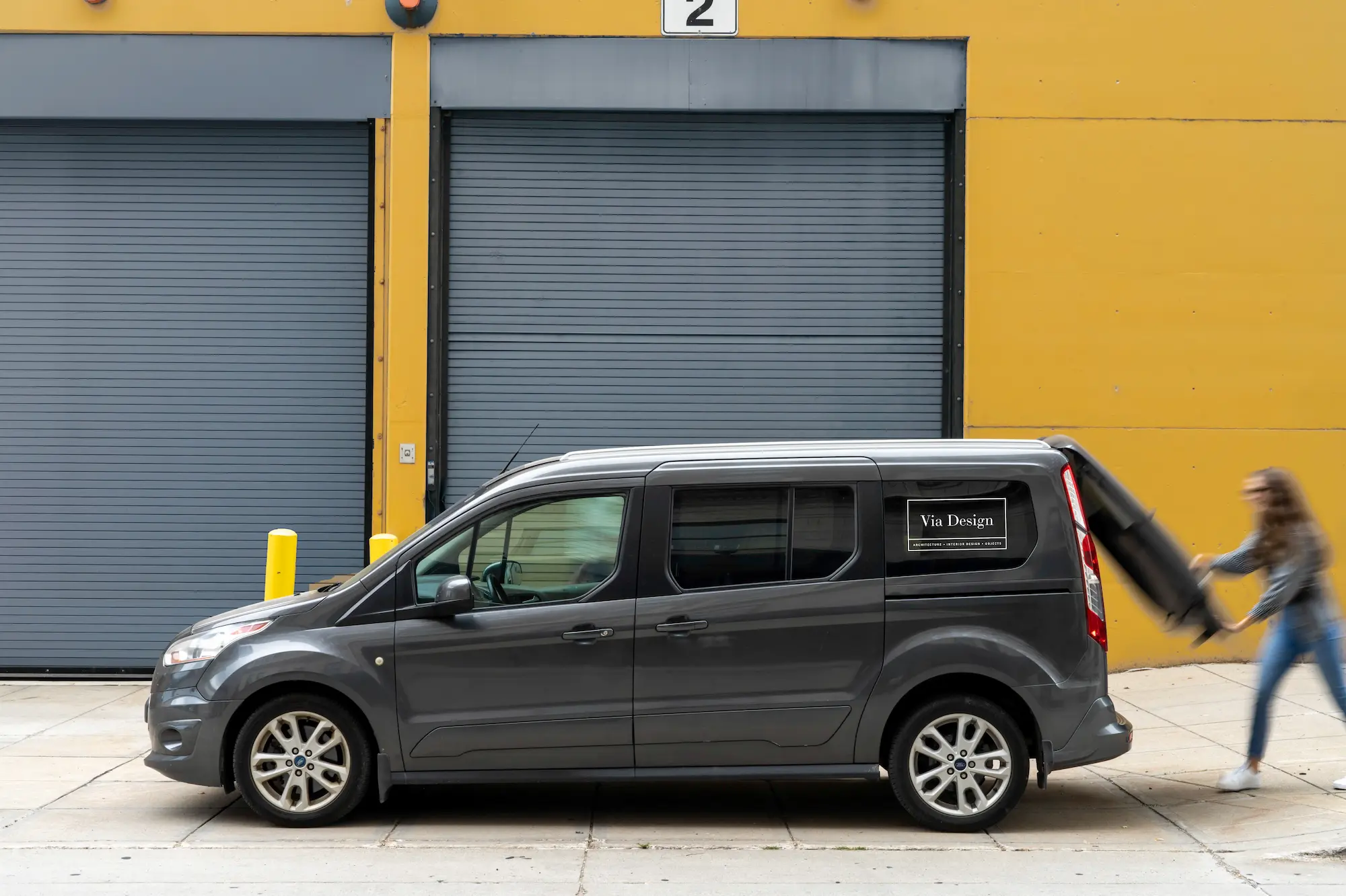 Gray van with Via Design logo parked by yellow wall, person loading large black item into back.