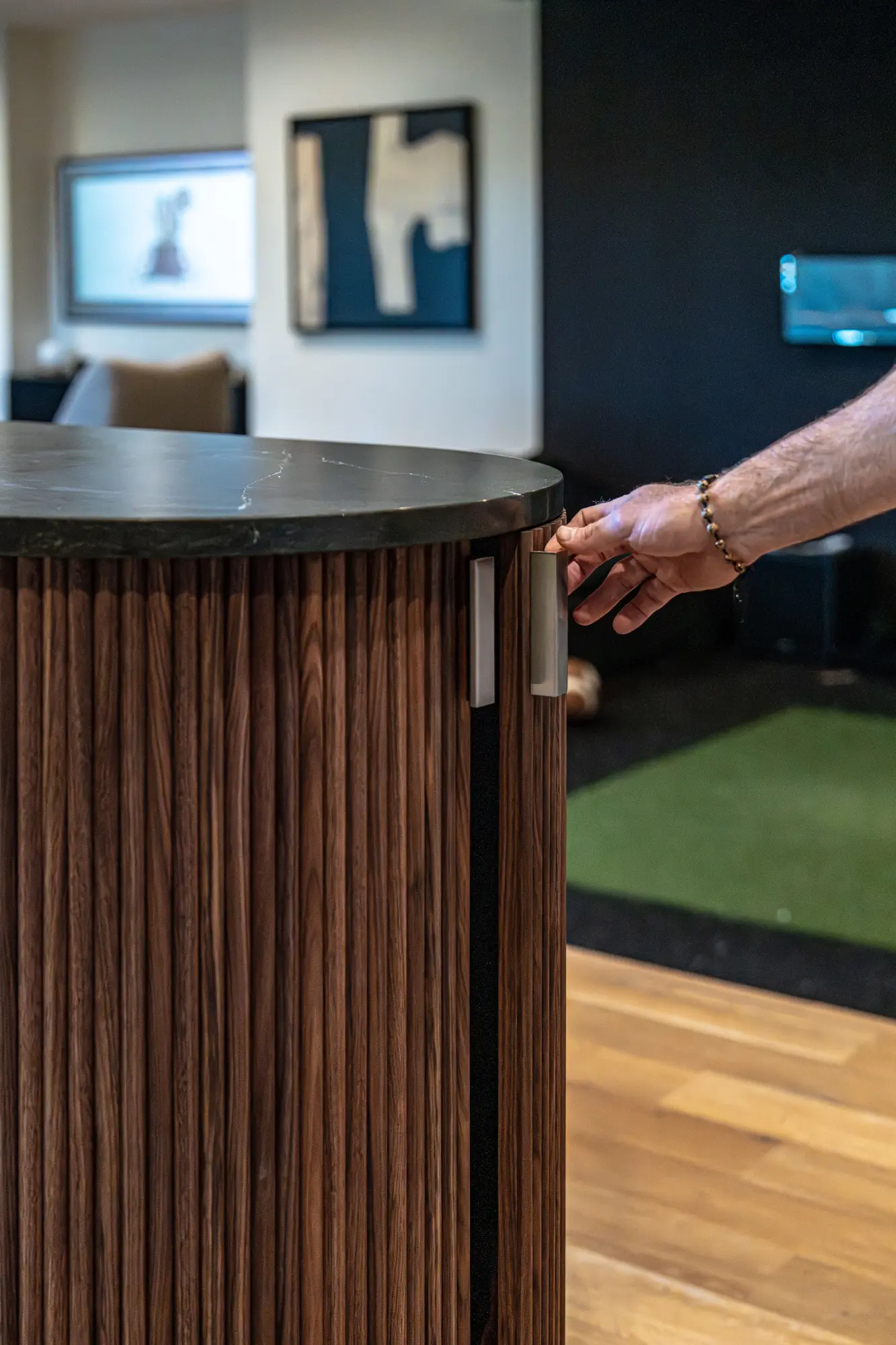 Close-up of a hand with a beaded bracelet reaching to open a dark wood cabinet door with vertical slats and a dark stone countertop in a modern interior.